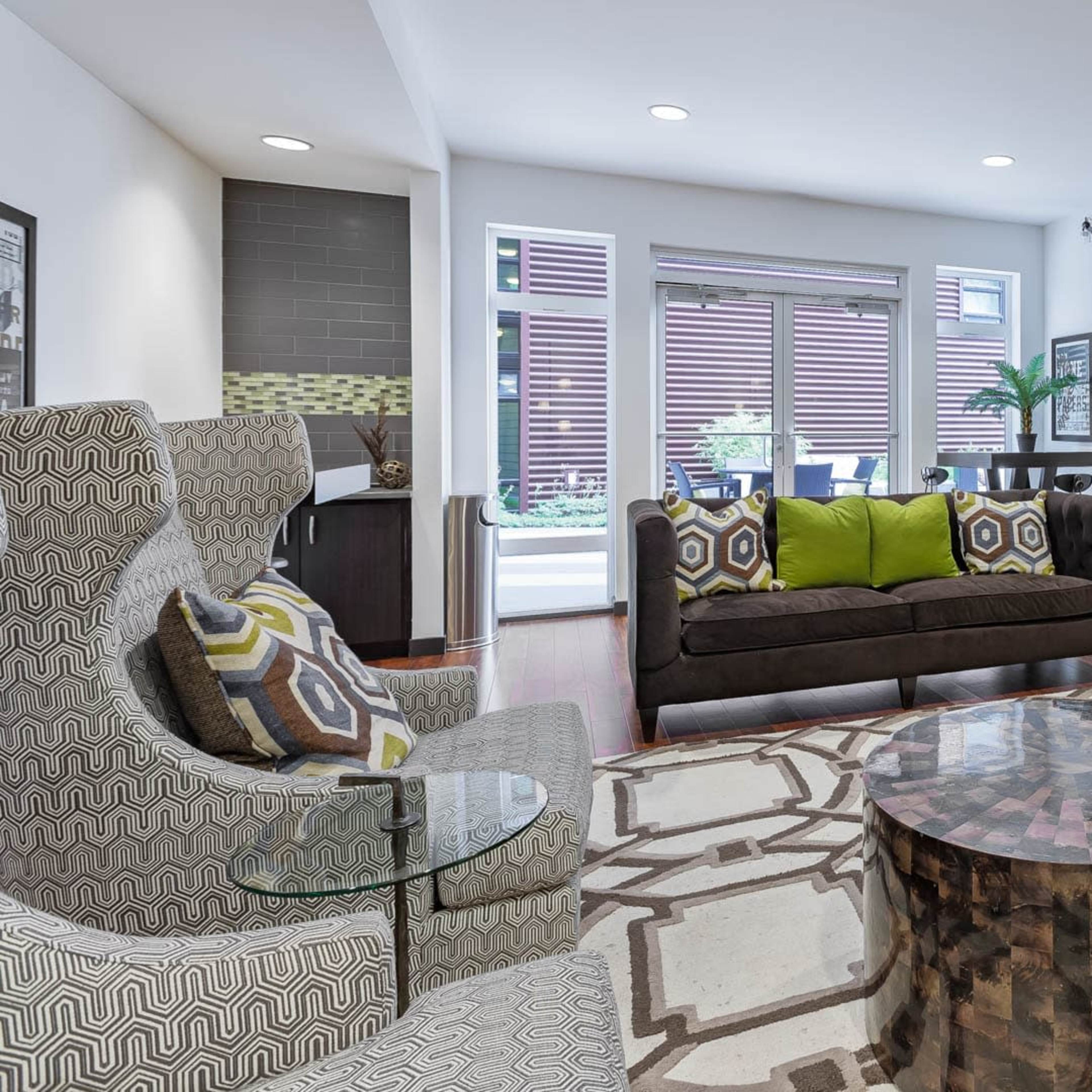 A modern living room features a patterned rug, a dark sofa with green and patterned pillows, two patterned chairs, a coffee table, a mounted television, and large windows with natural light.