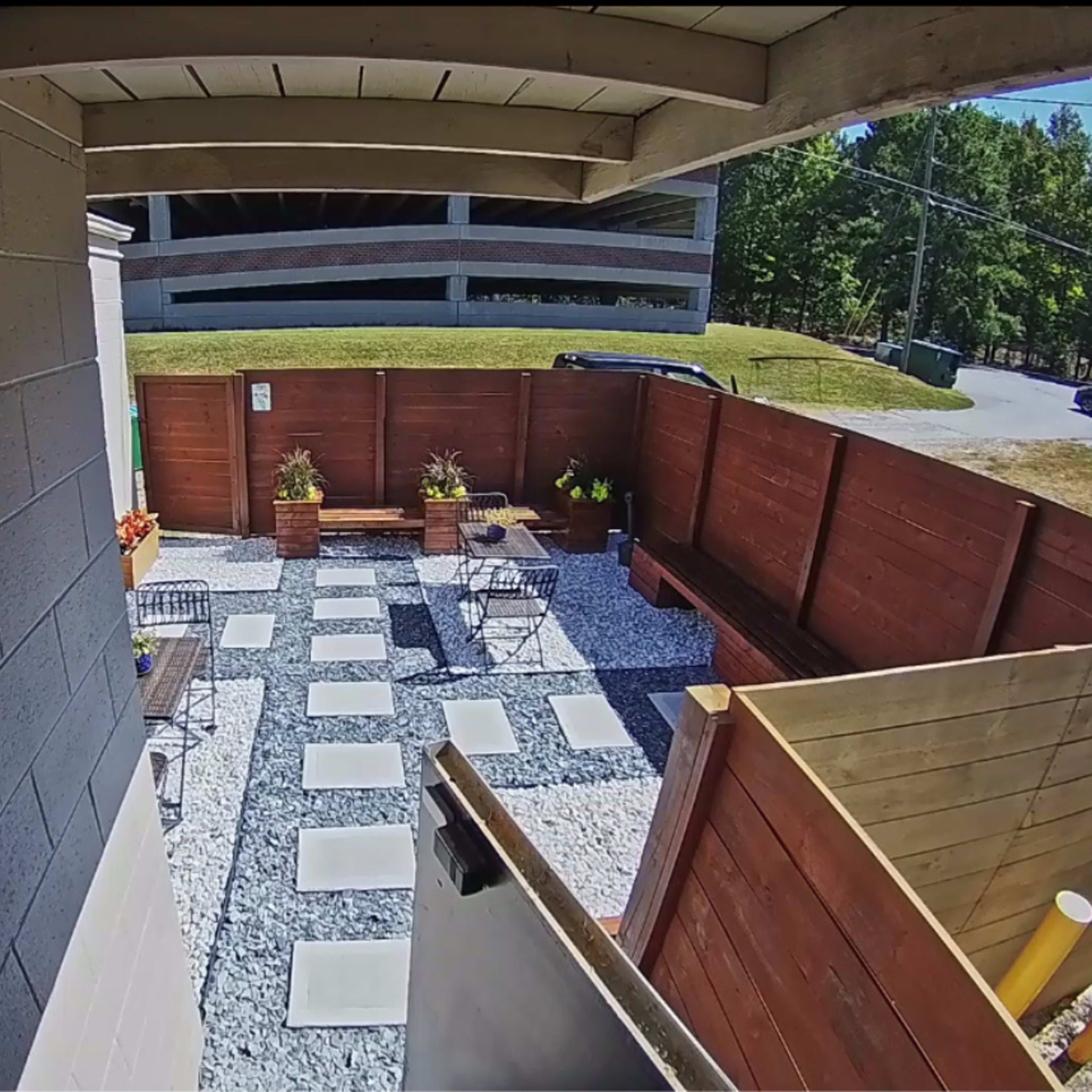 A patio area with gravel flooring, concrete stepping stones, and wooden fences, surrounded by potted plants and outdoor furniture.