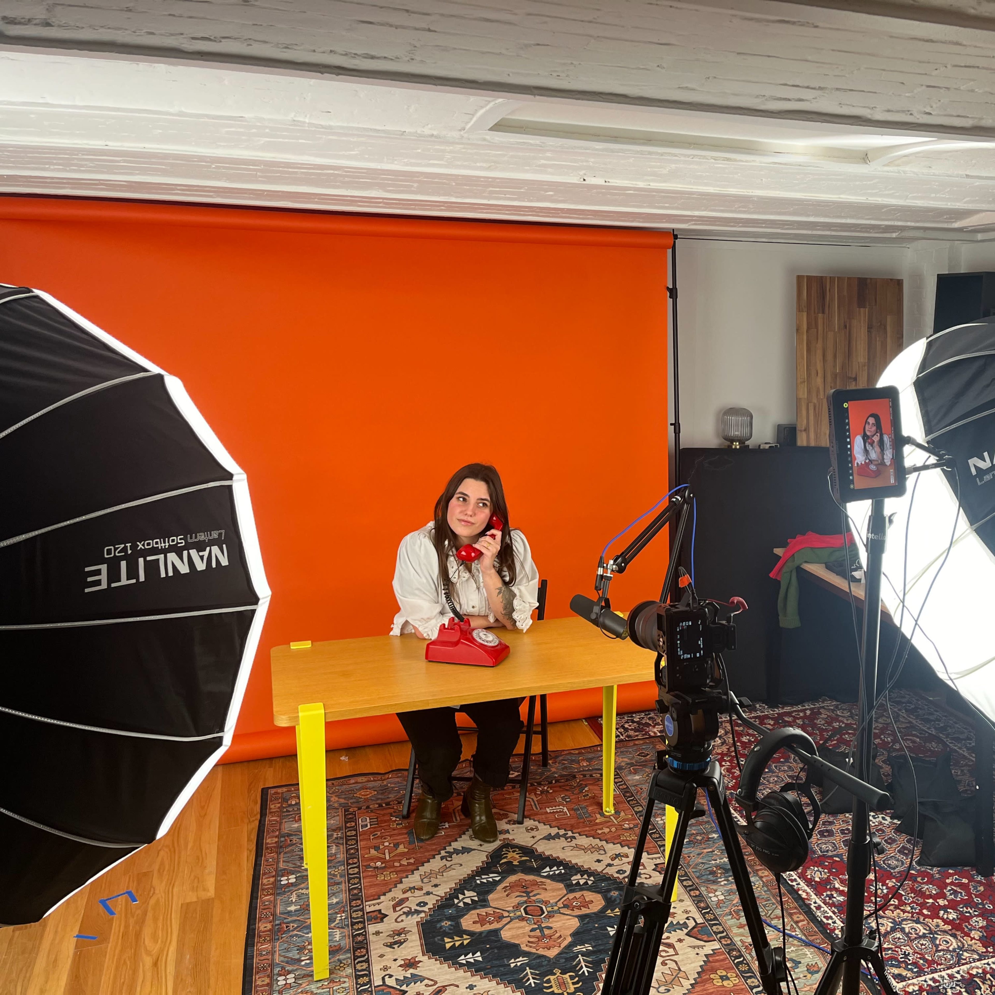 A woman sits at a table with a red purse in front of an orange backdrop, flanked by large softbox lights and a camera setup.