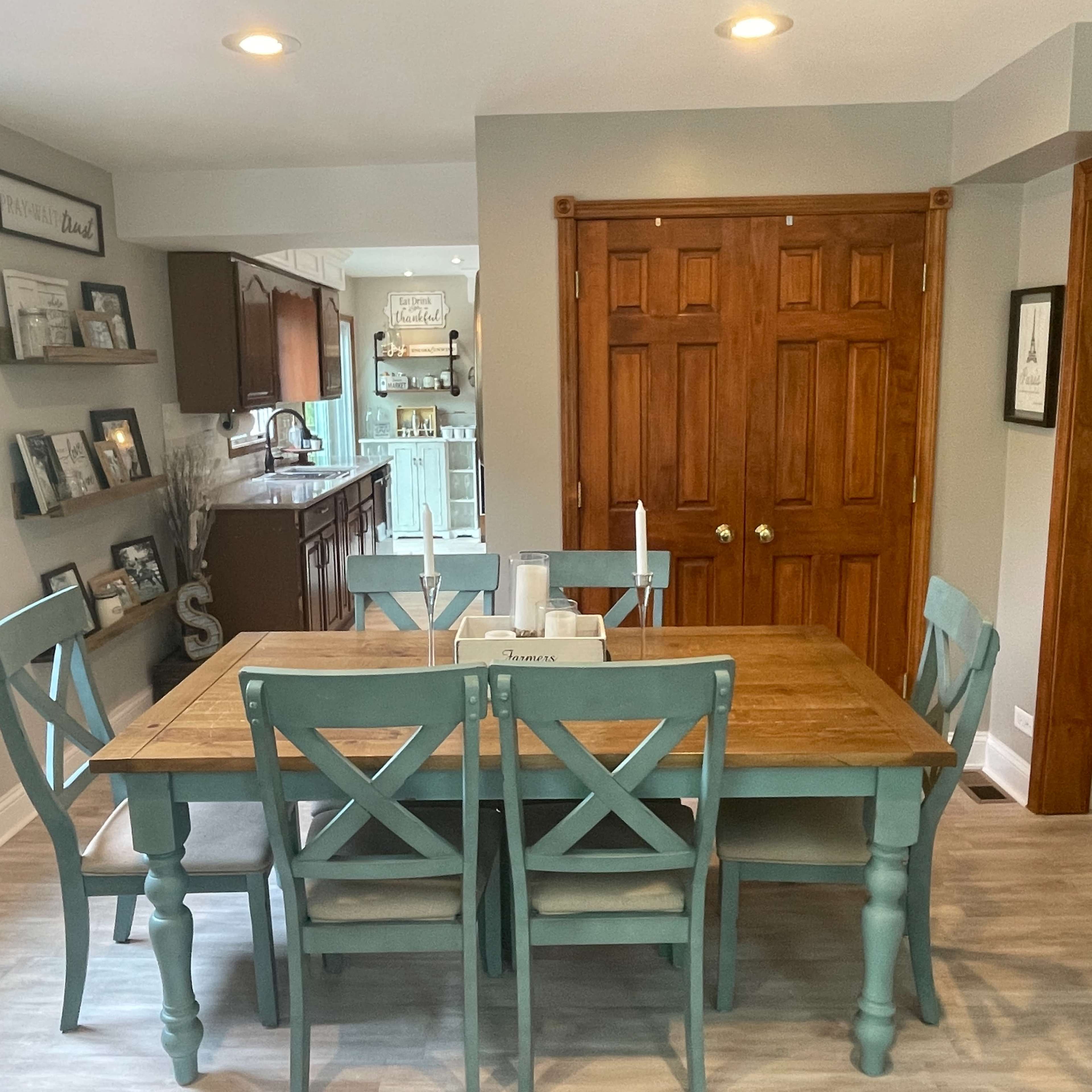 A dining room features a wooden table surrounded by four blue chairs, with a view of a kitchen in the background.