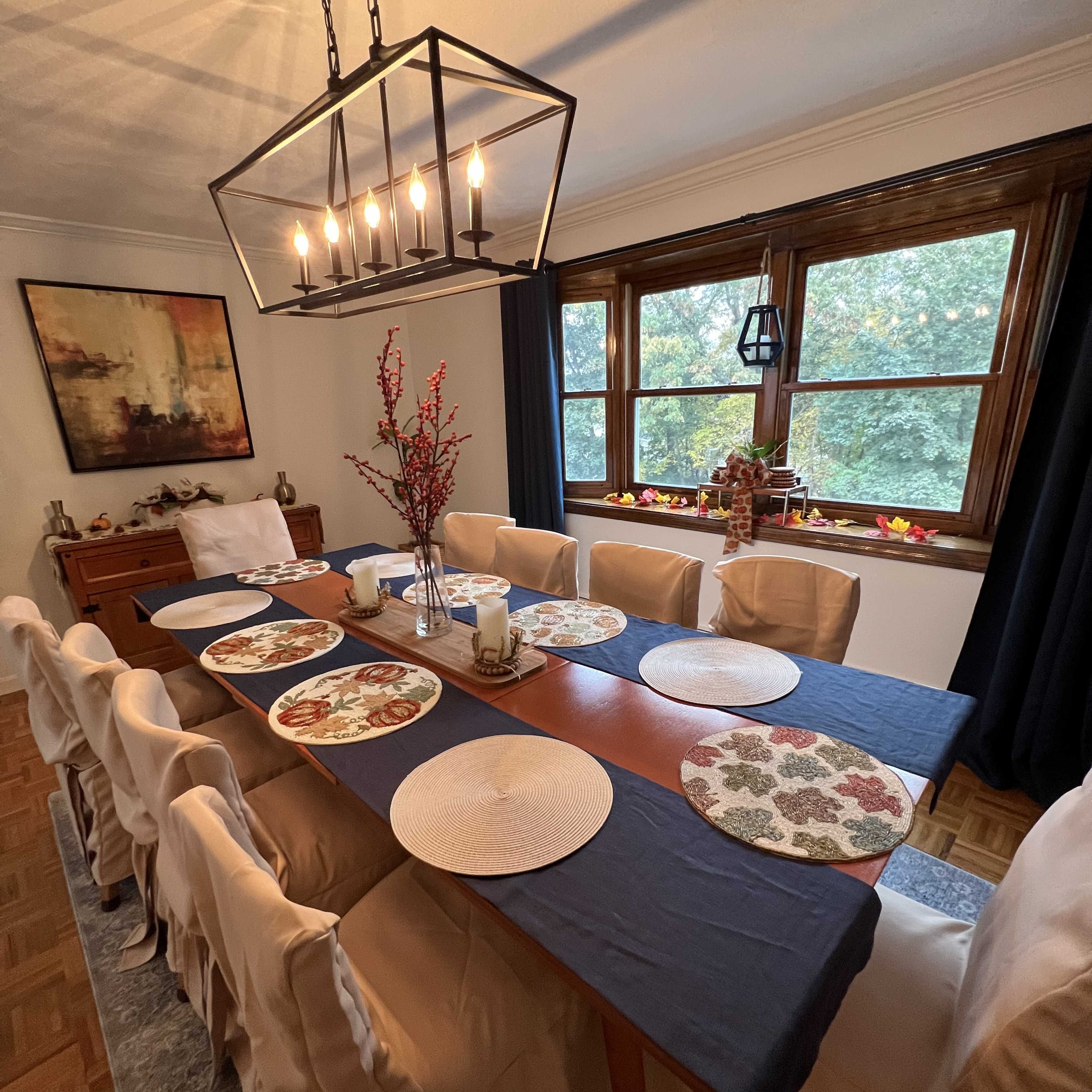A long dining table set with decorative placemats and a central floral arrangement is illuminated by a large chandelier in a well-lit room with large windows.