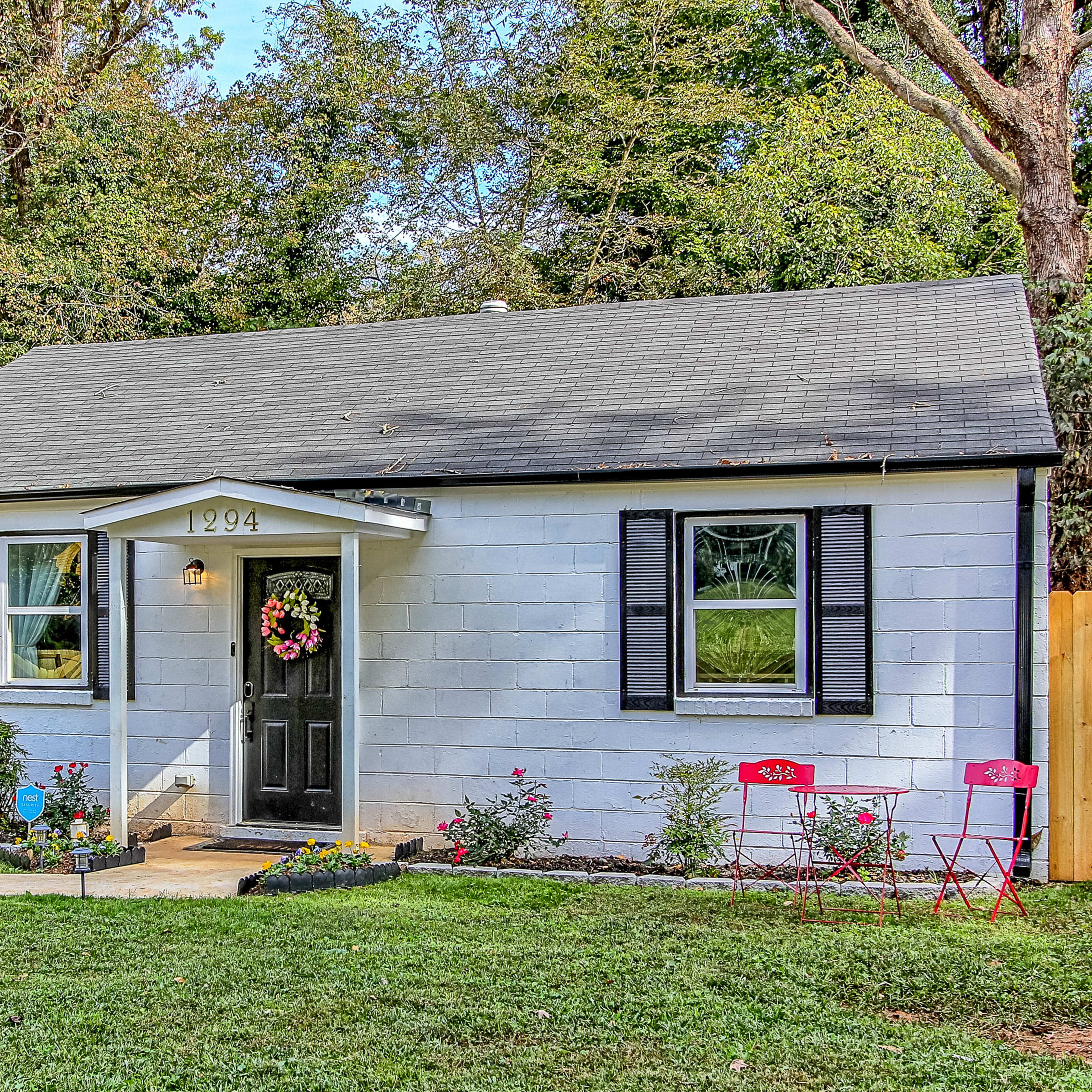 A small, single-story house with light blue siding and black shutters features a front porch with a decorative wreath and two red chairs on the lawn.