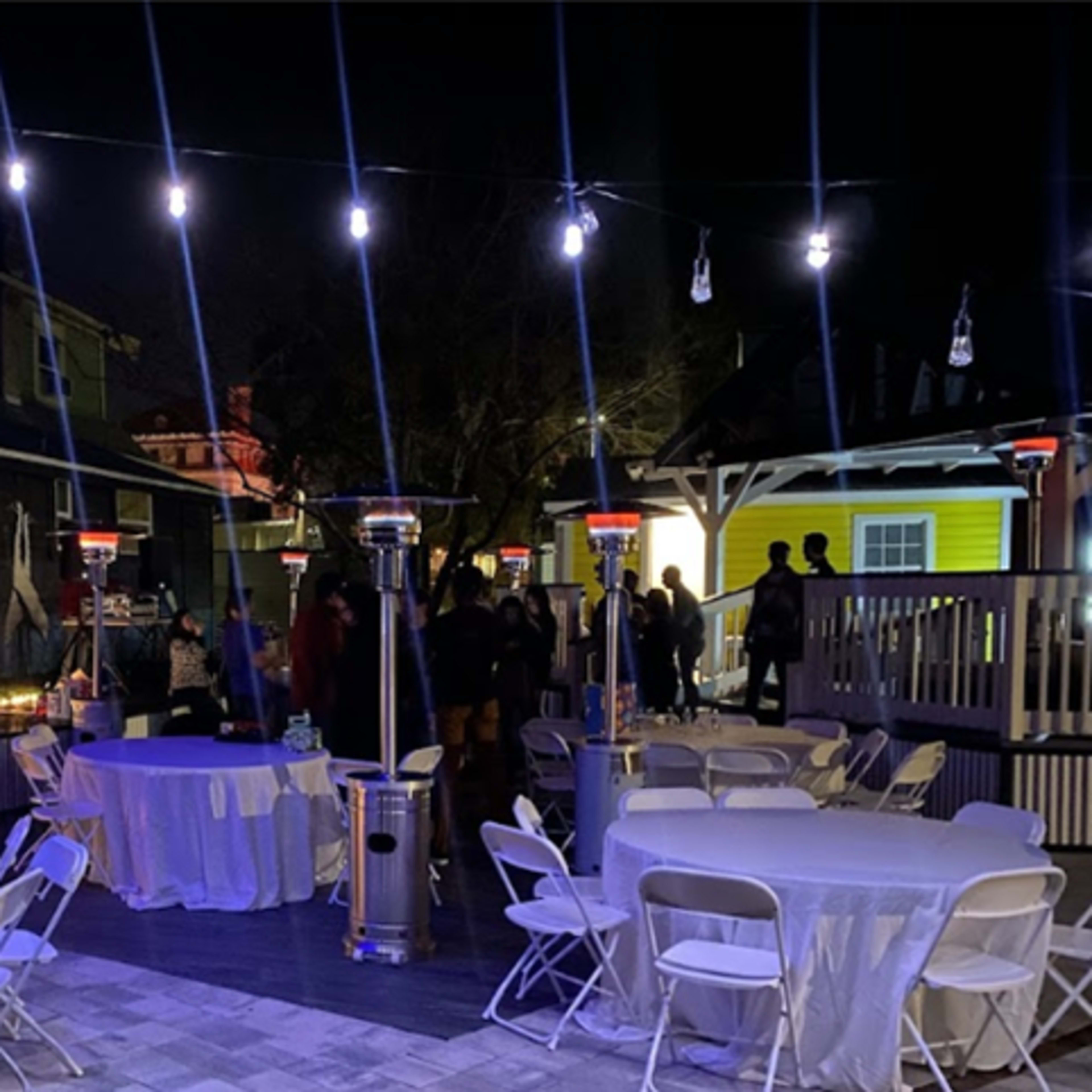A night event is set up outdoors with tables and heaters under string lights, while guests mingle near colorful buildings.