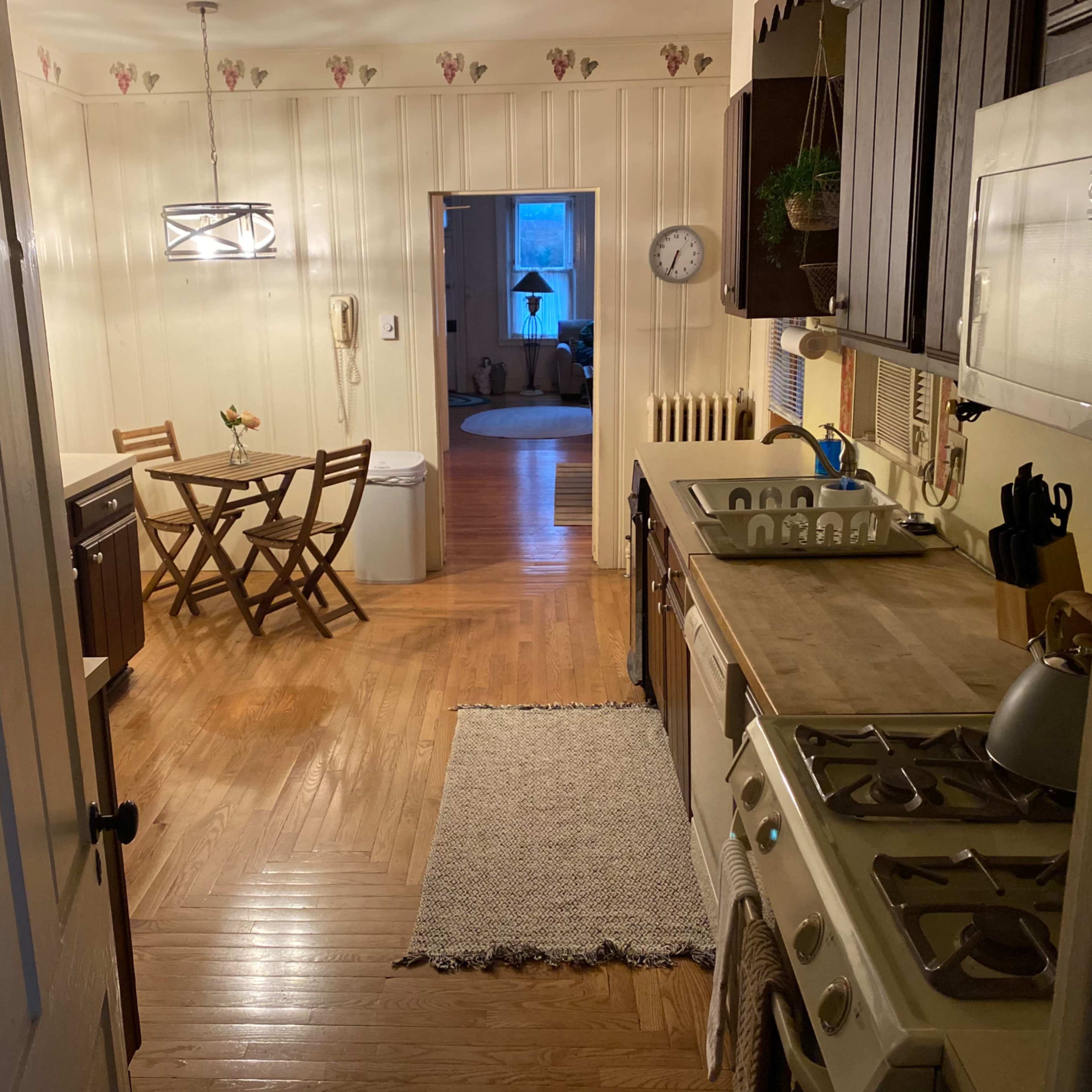 A kitchen with wooden cabinets and a stove leads into a dining area featuring a table and chairs in a well-lit space.