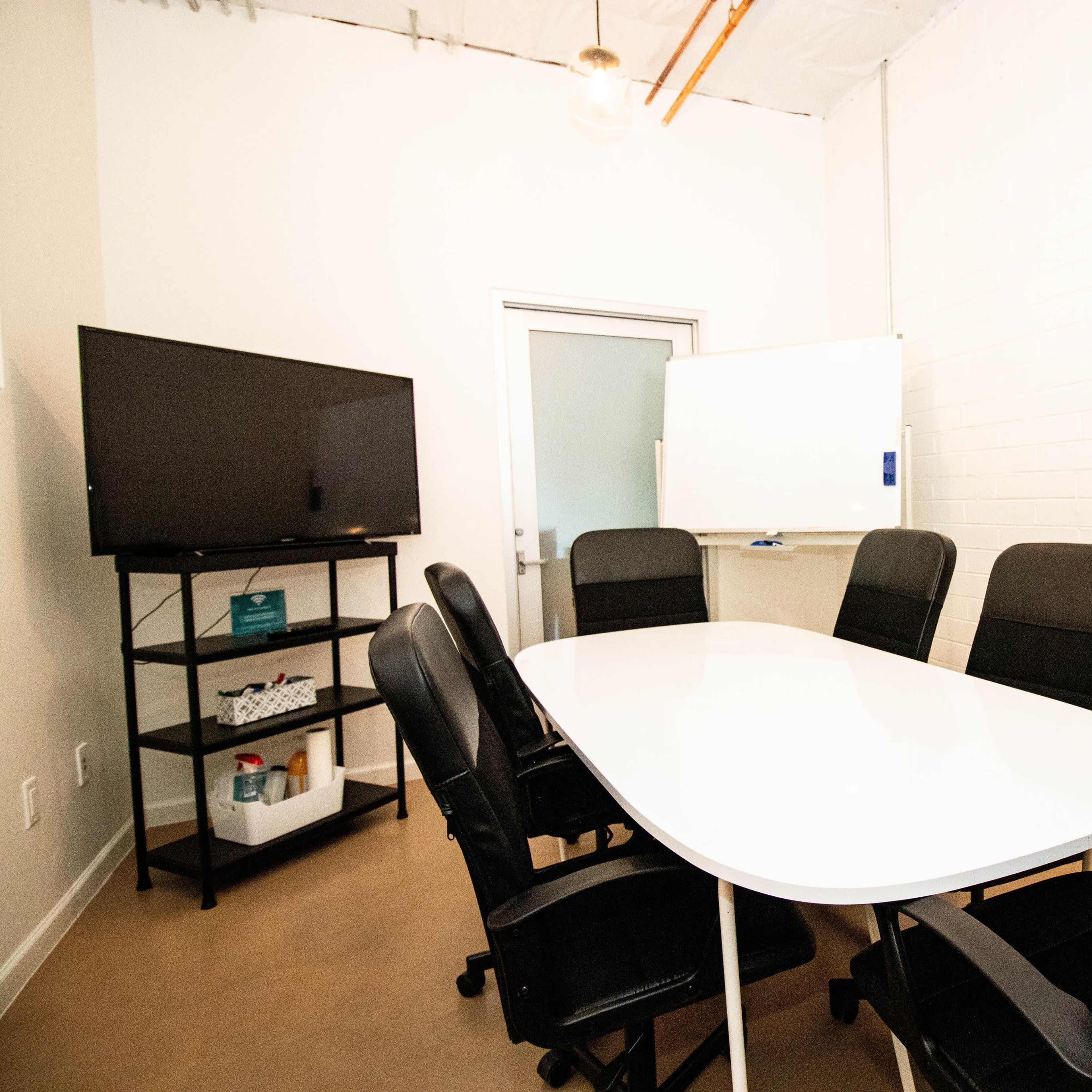 A conference room features a white oval table surrounded by six black ergonomic chairs, with a television and a whiteboard on the walls.