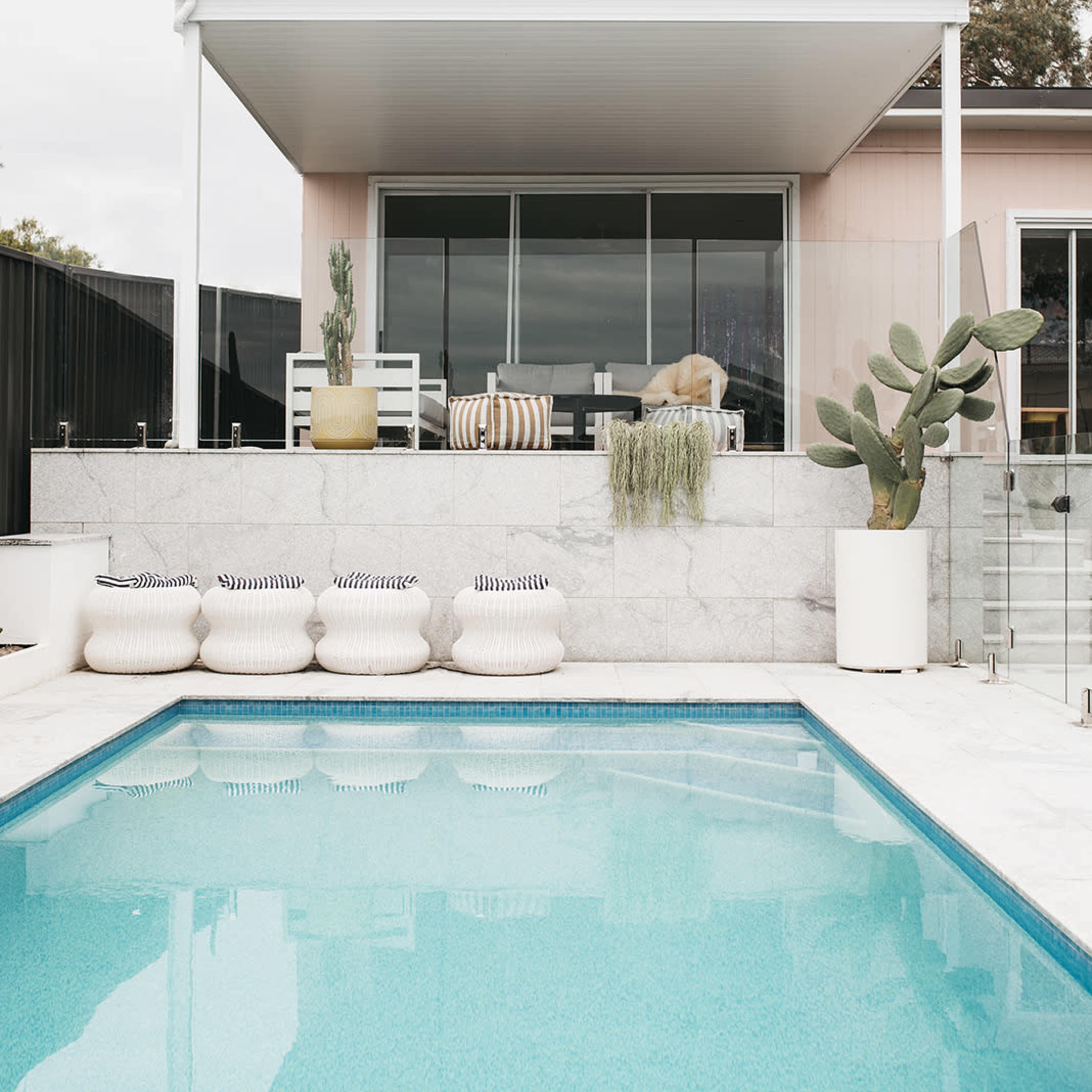A modern outdoor space features a swimming pool in the foreground, with a seating area and potted plants visible behind a glass wall.