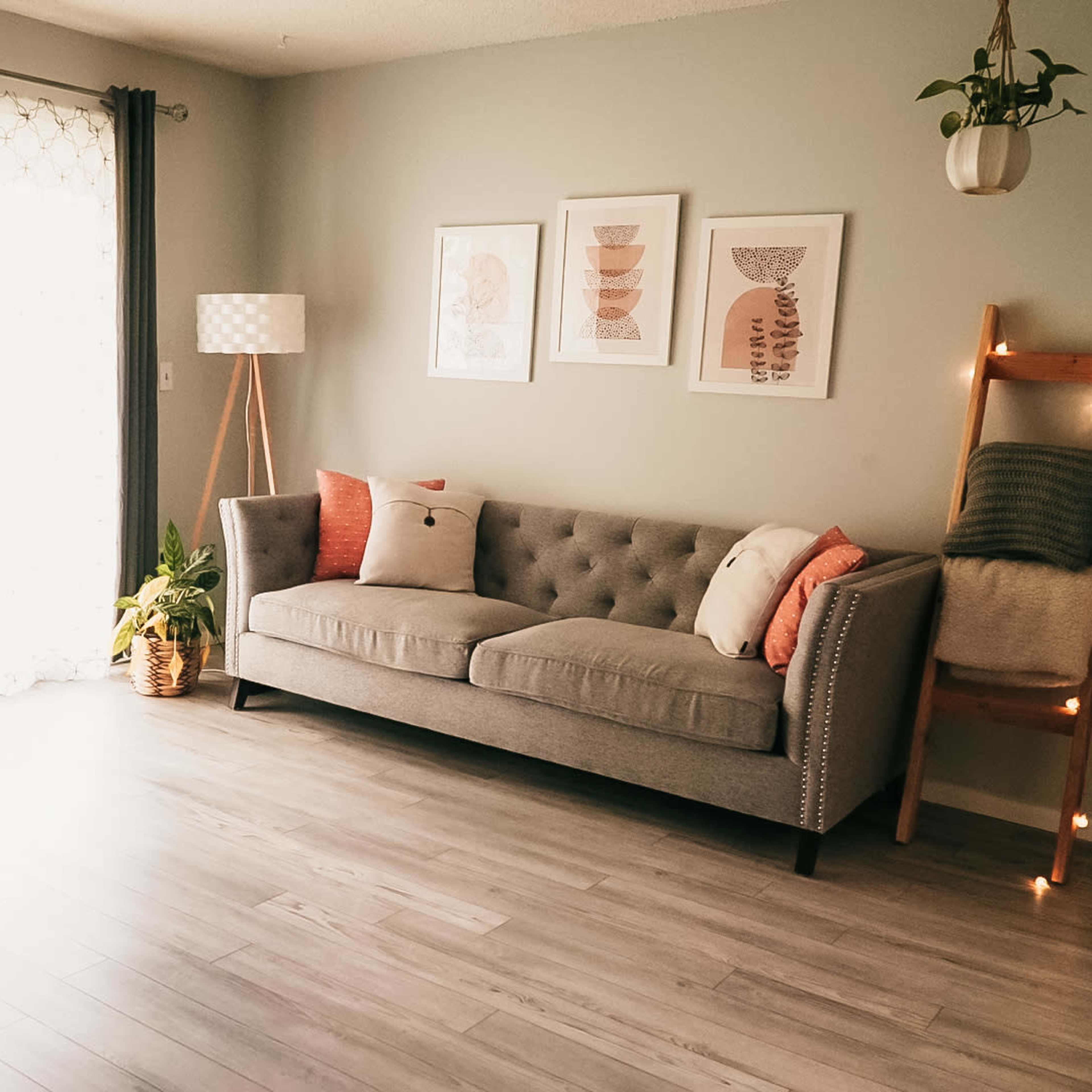 A gray tufted sofa with decorative cushions is positioned in a well-lit room featuring framed artwork, a floor lamp, and a wooden ladder displaying a blanket.
