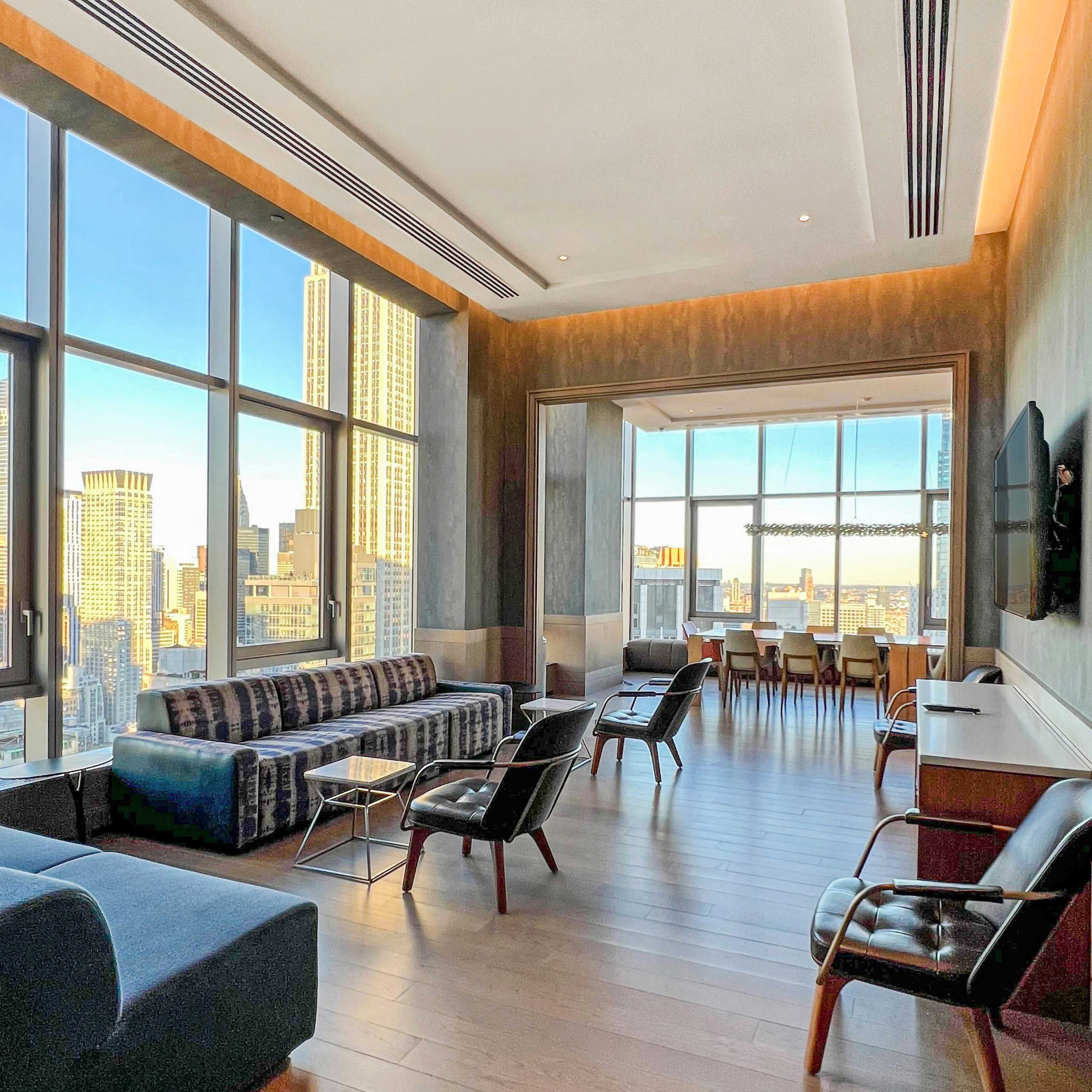 A modern lounge area in a high-rise building with large windows offering a city view, featuring contemporary furniture including sofas and chairs.