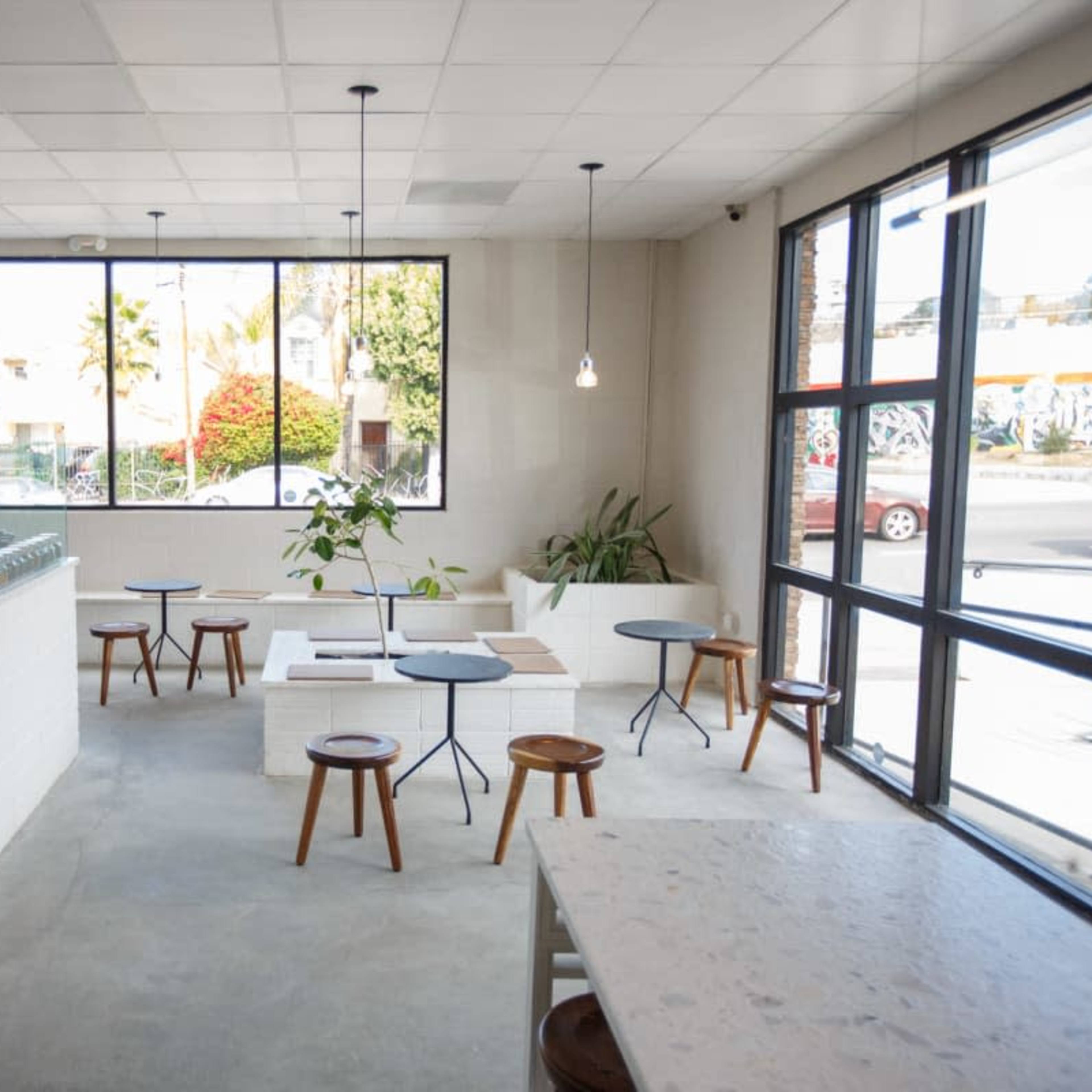 A modern café interior with a spacious layout, featuring a counter on the left, small tables with chairs, and large windows overlooking the street.