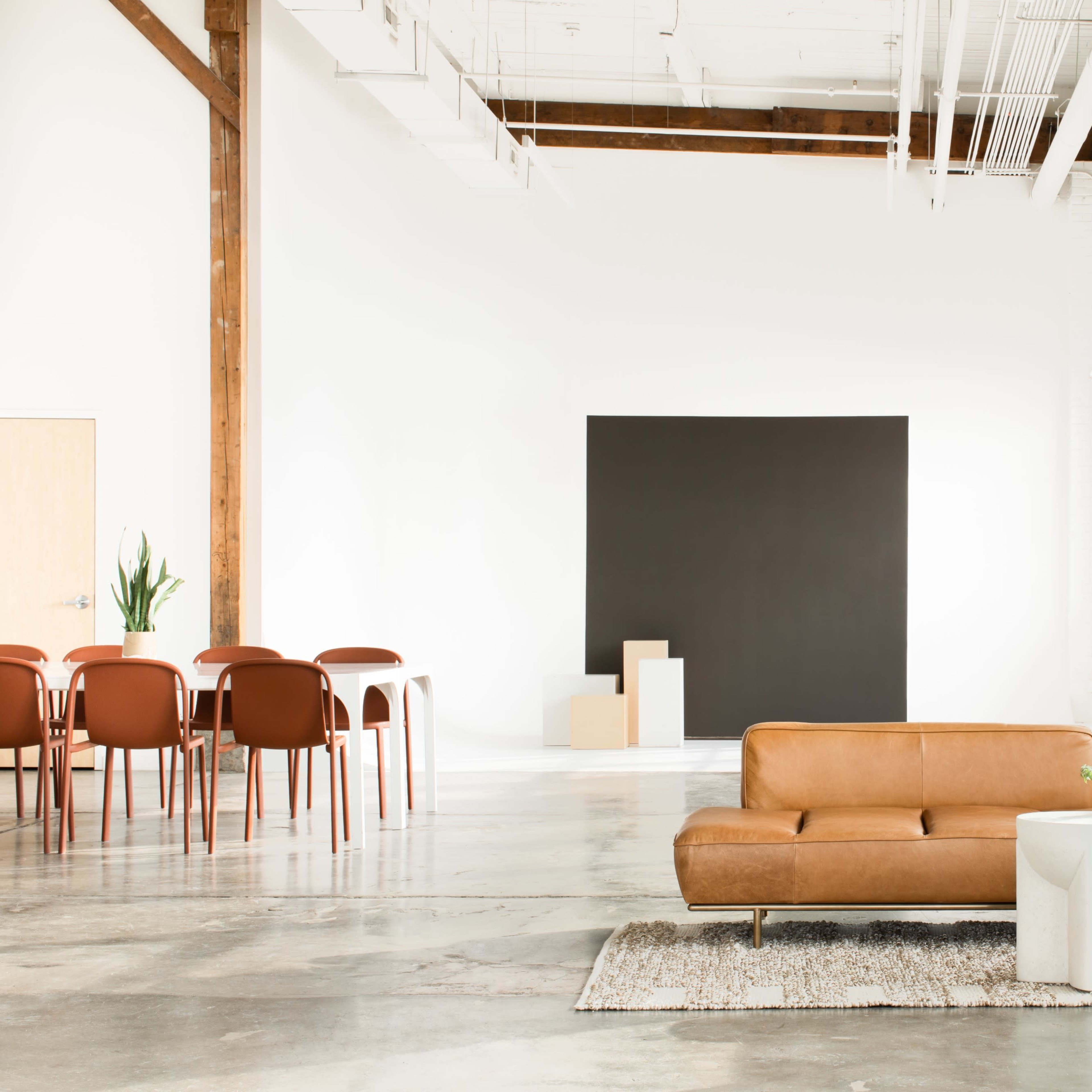 A leather sofa sits on a rug in a spacious room with large windows, a black wall, and a dining area featuring wooden chairs and tables.
