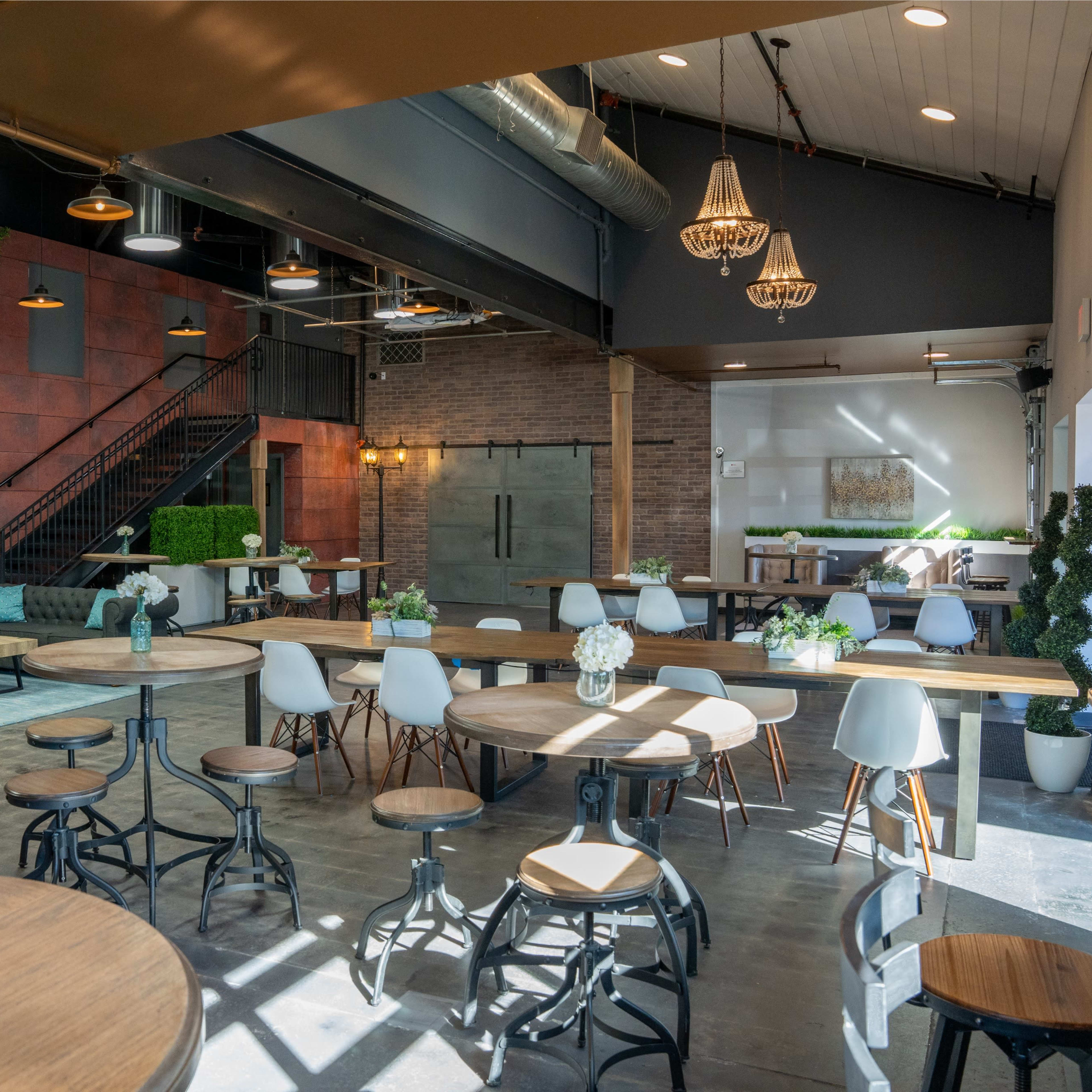 A spacious indoor area with a combination of wooden tables, chairs, and plants, featuring a staircase in the background and large windows that allow natural light to enter.
