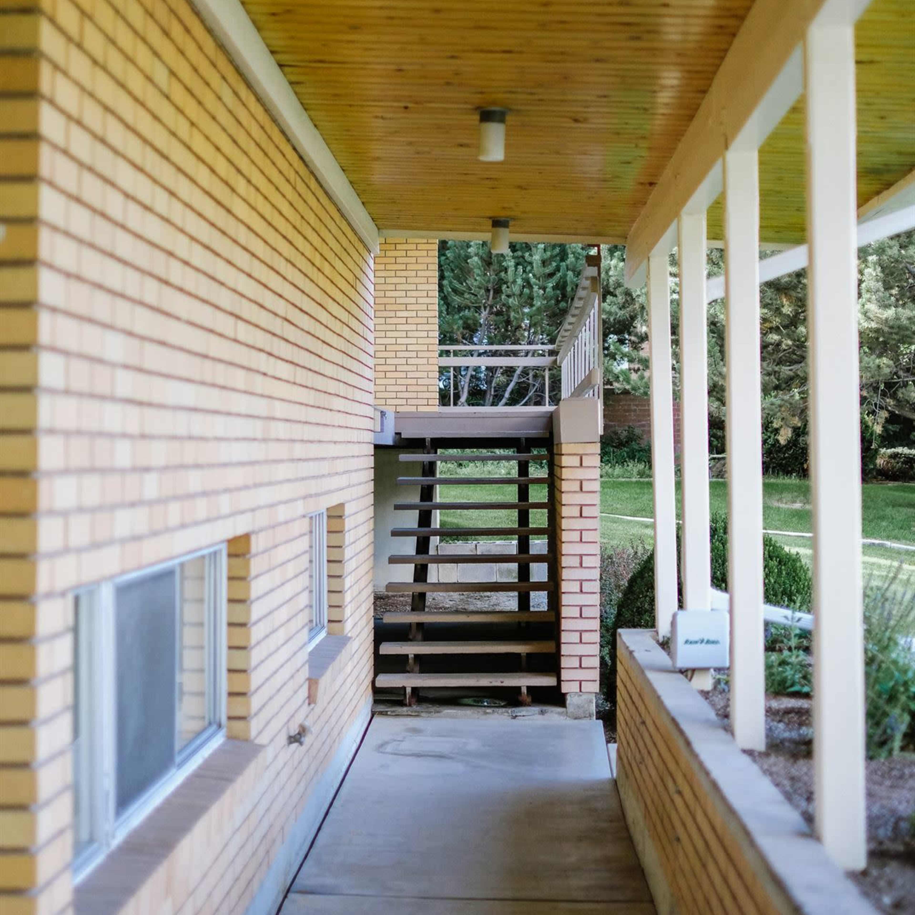 A covered walkway leads to a set of wooden stairs beside a brick building.