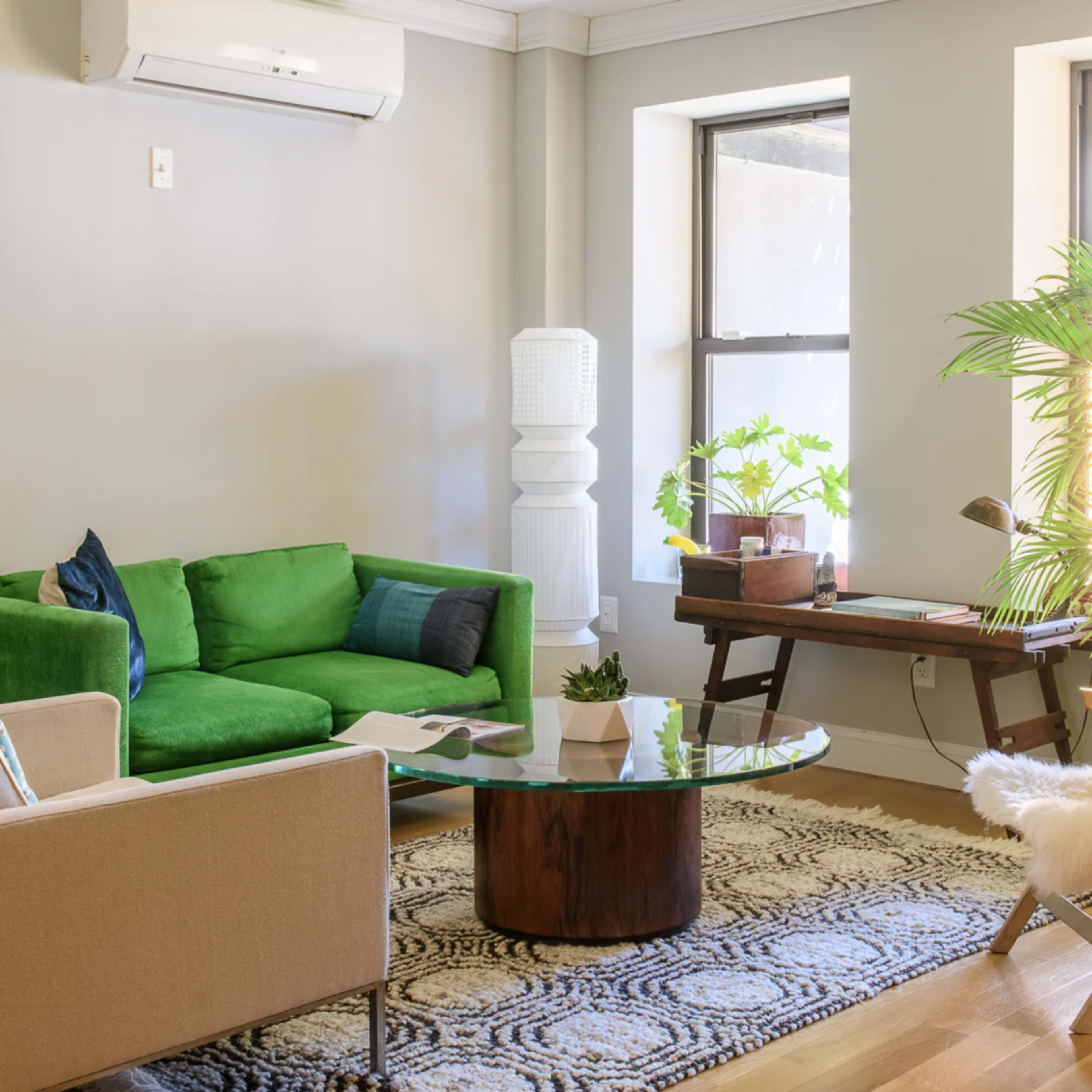 A bright living room featuring a green sofa, a round glass coffee table, a workspace with a wooden desk, and plants by a large window.