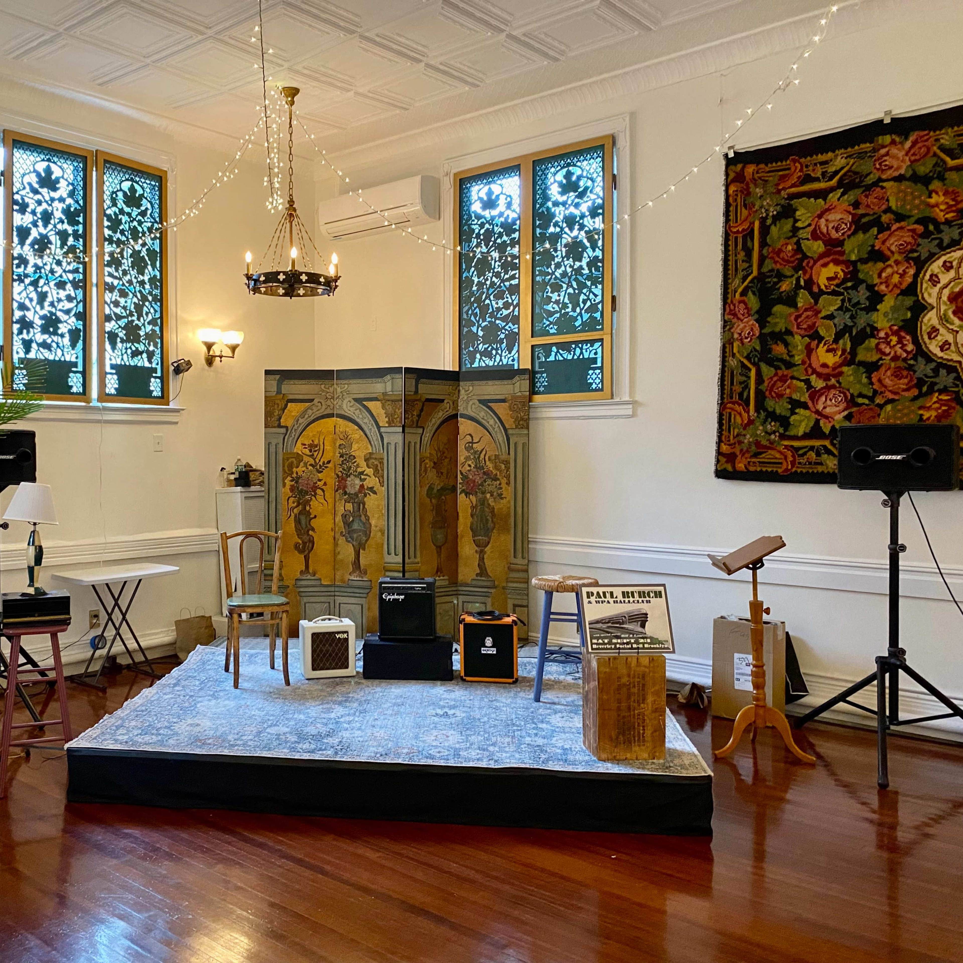 A performance stage is set up in a room with decorative windows, featuring a carpet, microphones, and various music equipment.