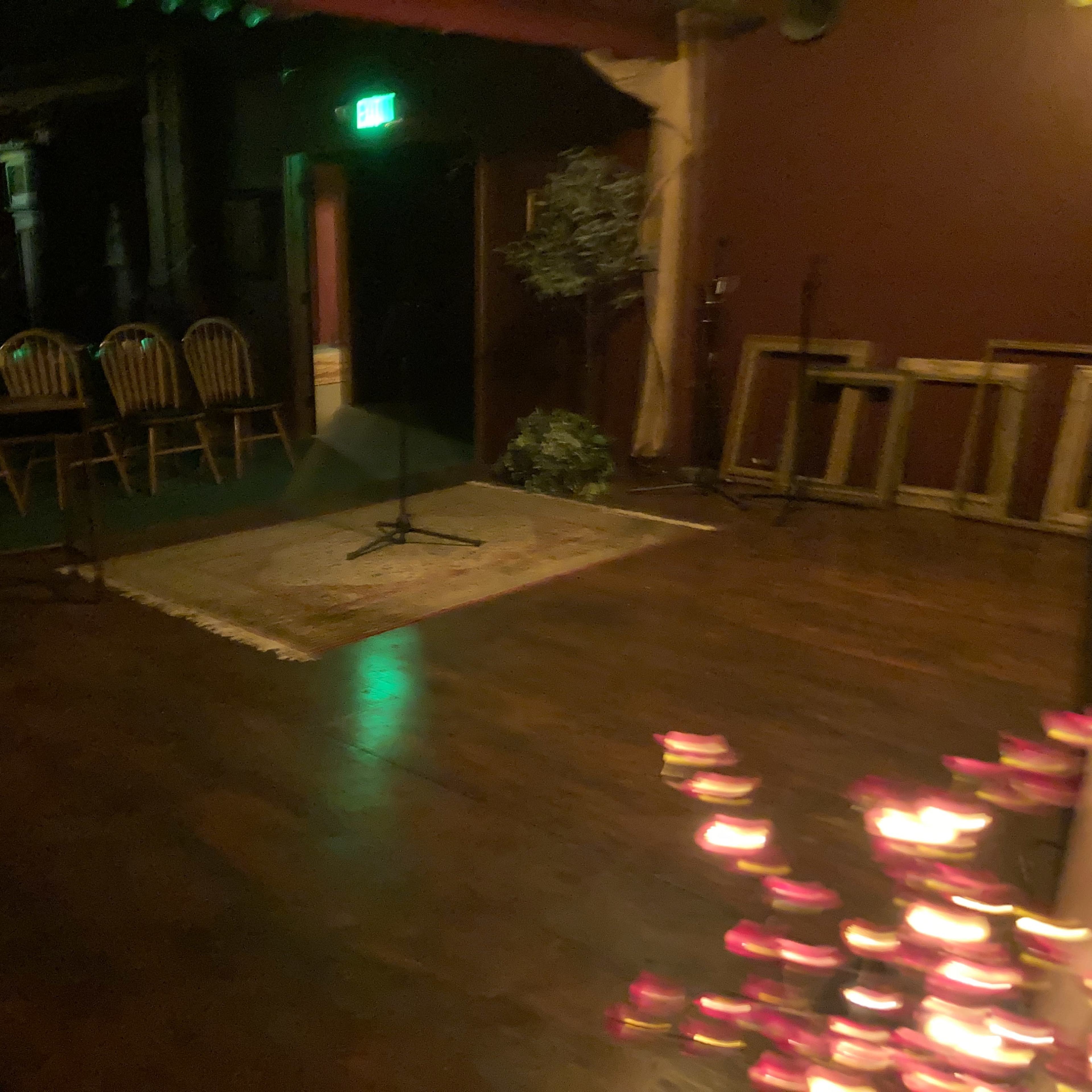 A dimly lit performance space features a wooden floor, a small rug, and several chairs arranged around a table.