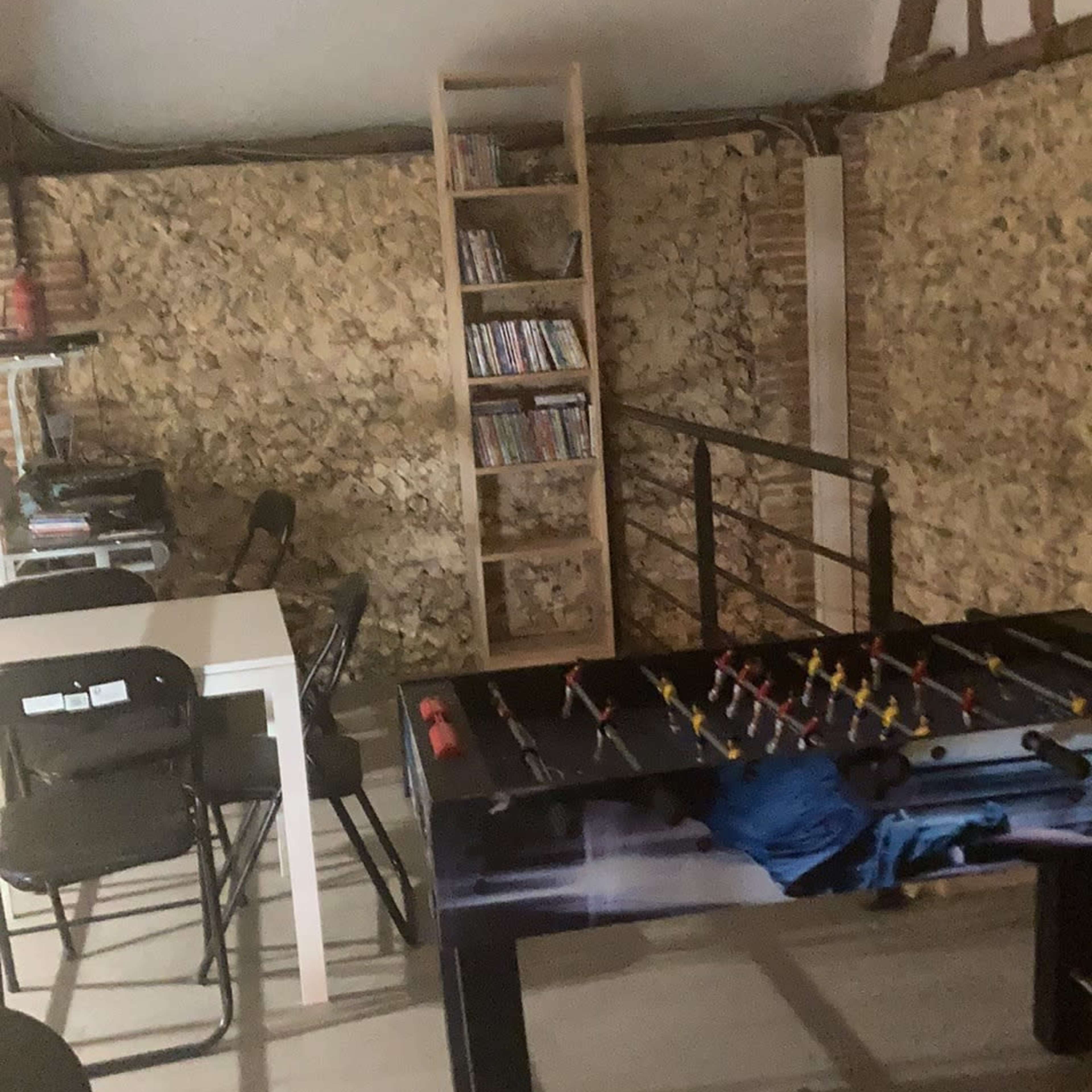 A room with a foosball table, a white table, black chairs, and a bookshelf filled with DVDs against a rustic stone wall.