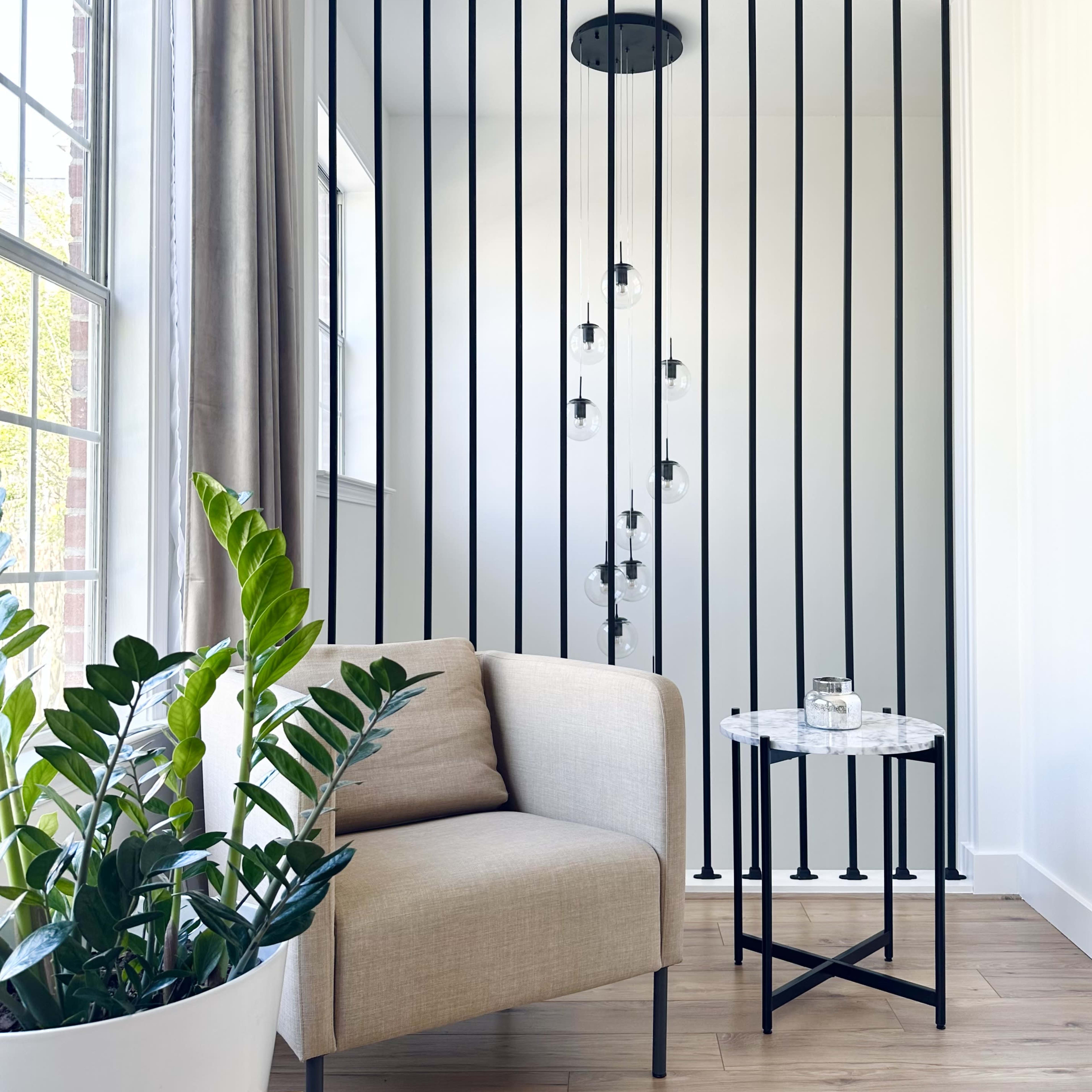 A beige armchair and side table are positioned beside a planter with a plant, separated from a well-lit area by vertical black bars and hanging light fixtures.