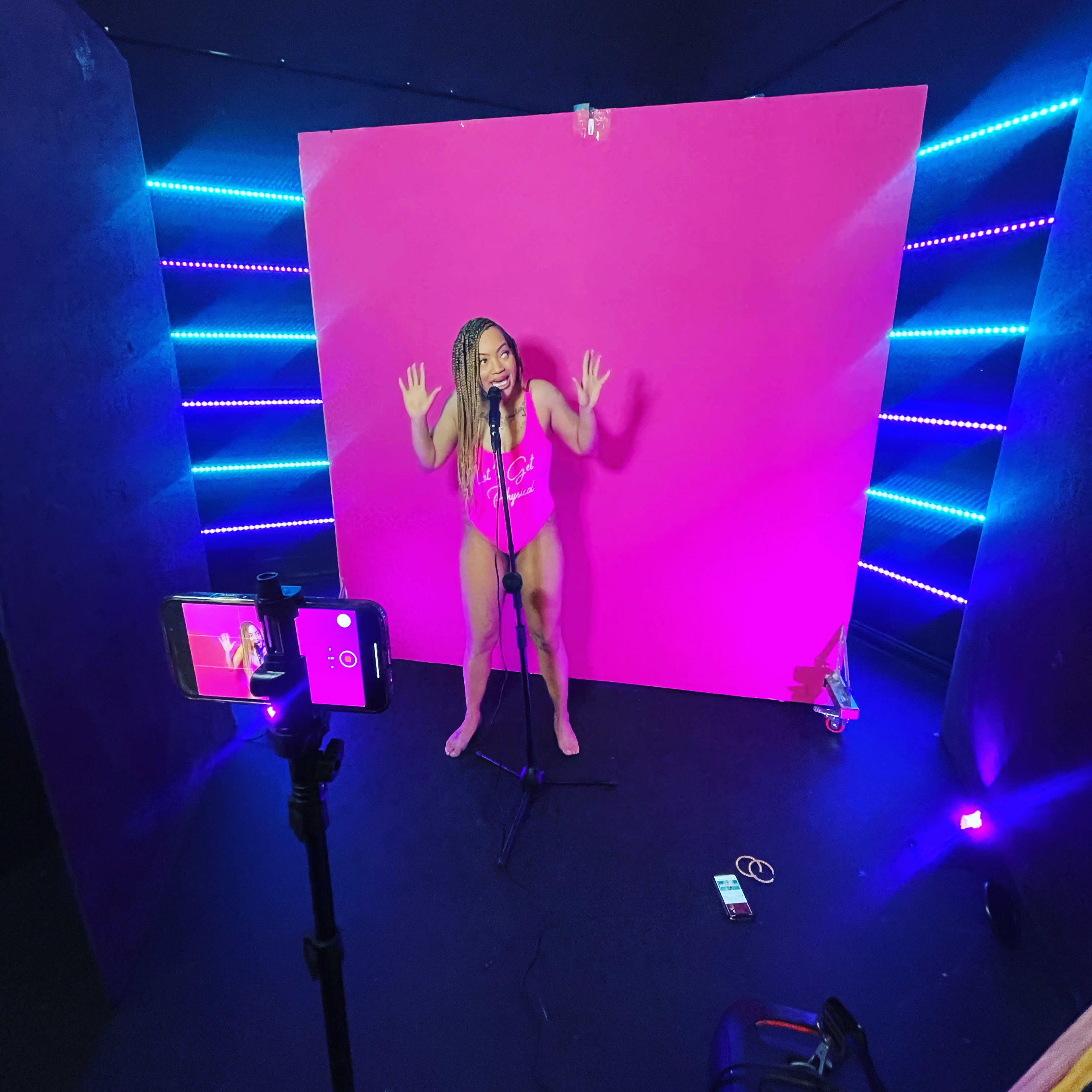A performer stands in front of a bright pink backdrop, holding a microphone, with colored lights illuminating the surrounding space.