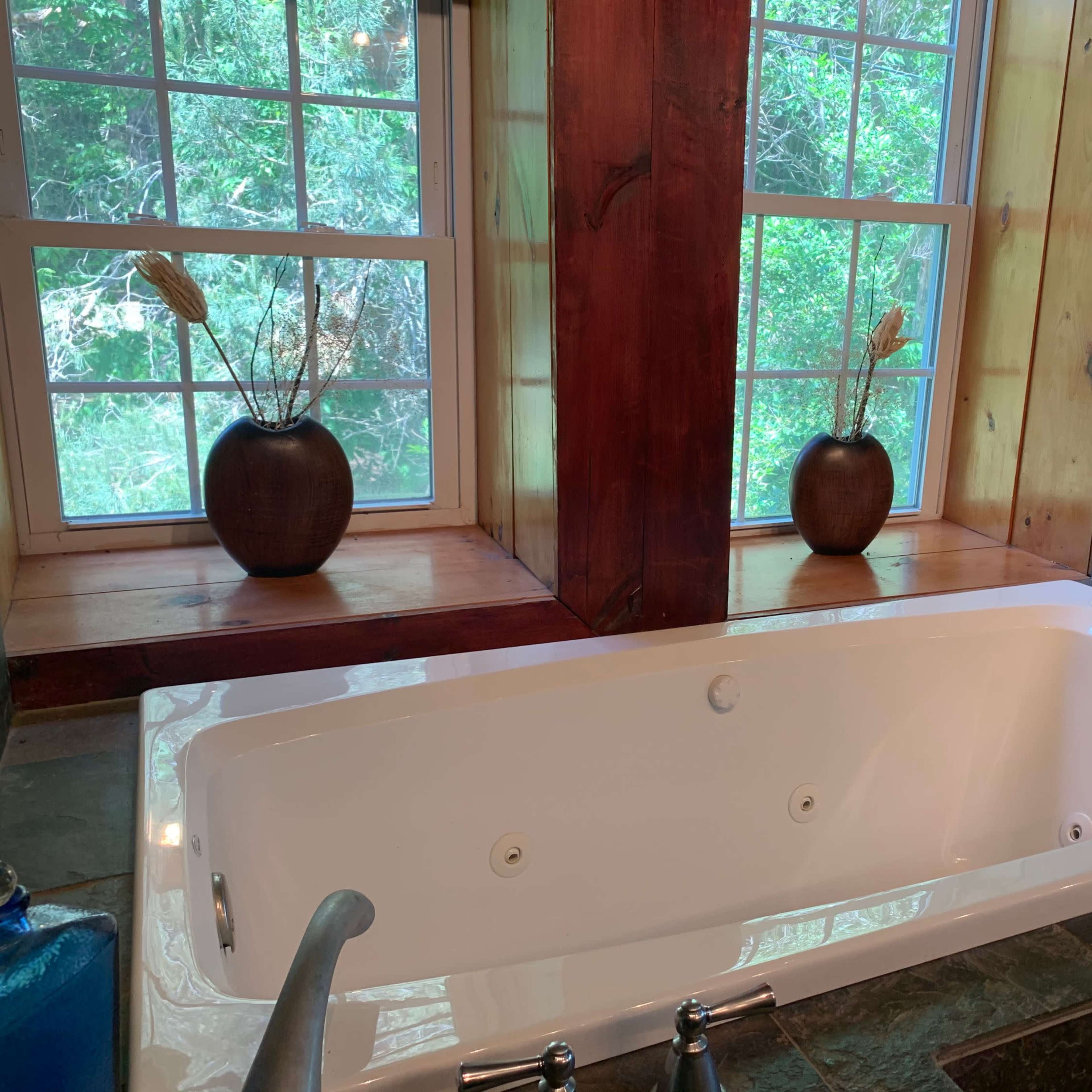 A white bathtub is positioned beside two windows framed with wooden panels, which overlook greenery outside.