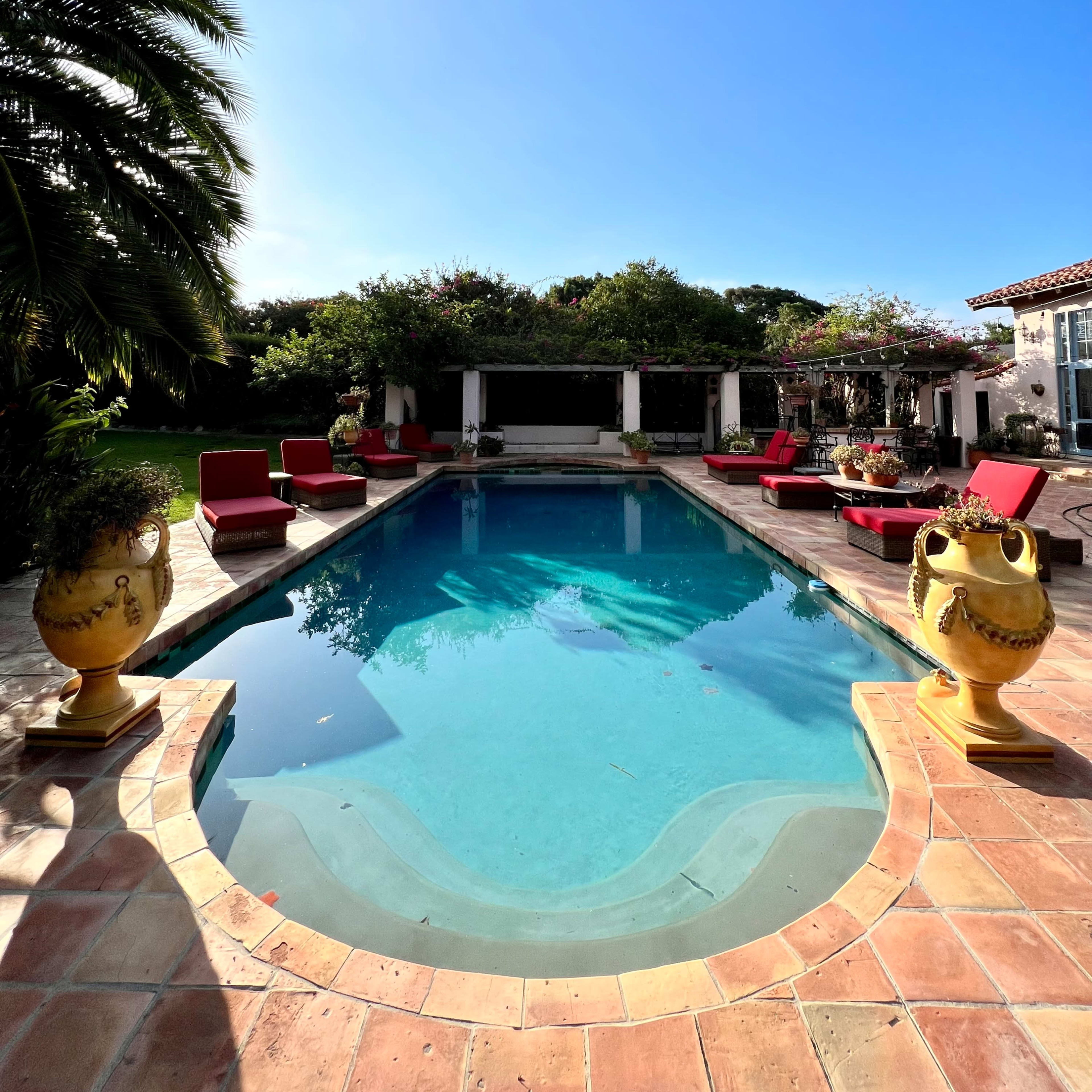 A clear swimming pool is surrounded by terracotta tiles and red lounge chairs, set against a backdrop of lush greenery and a bright blue sky.