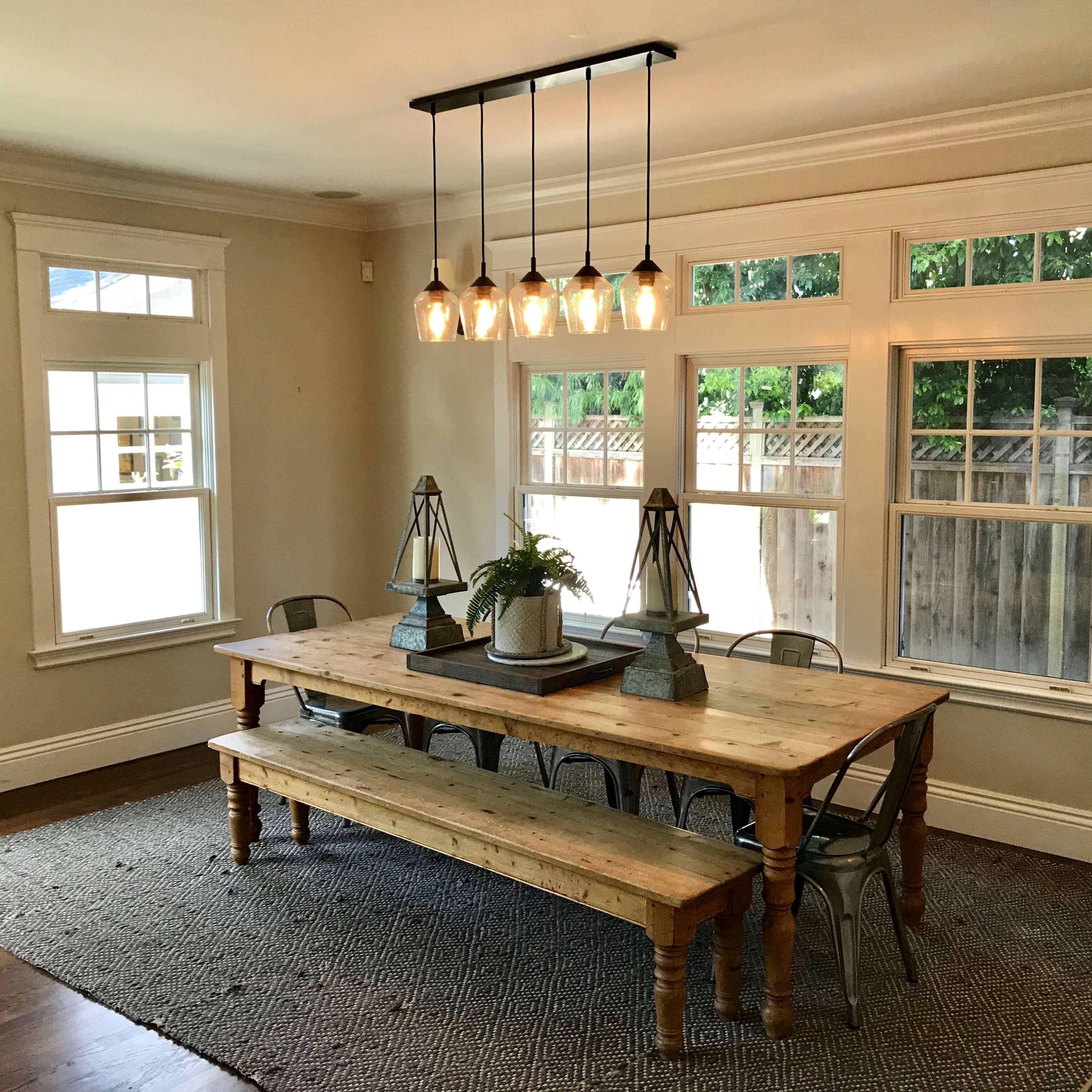 A large wooden dining table is centered in a well-lit room with multiple windows and a pendant light overhead, surrounded by metal chairs and decorative elements.