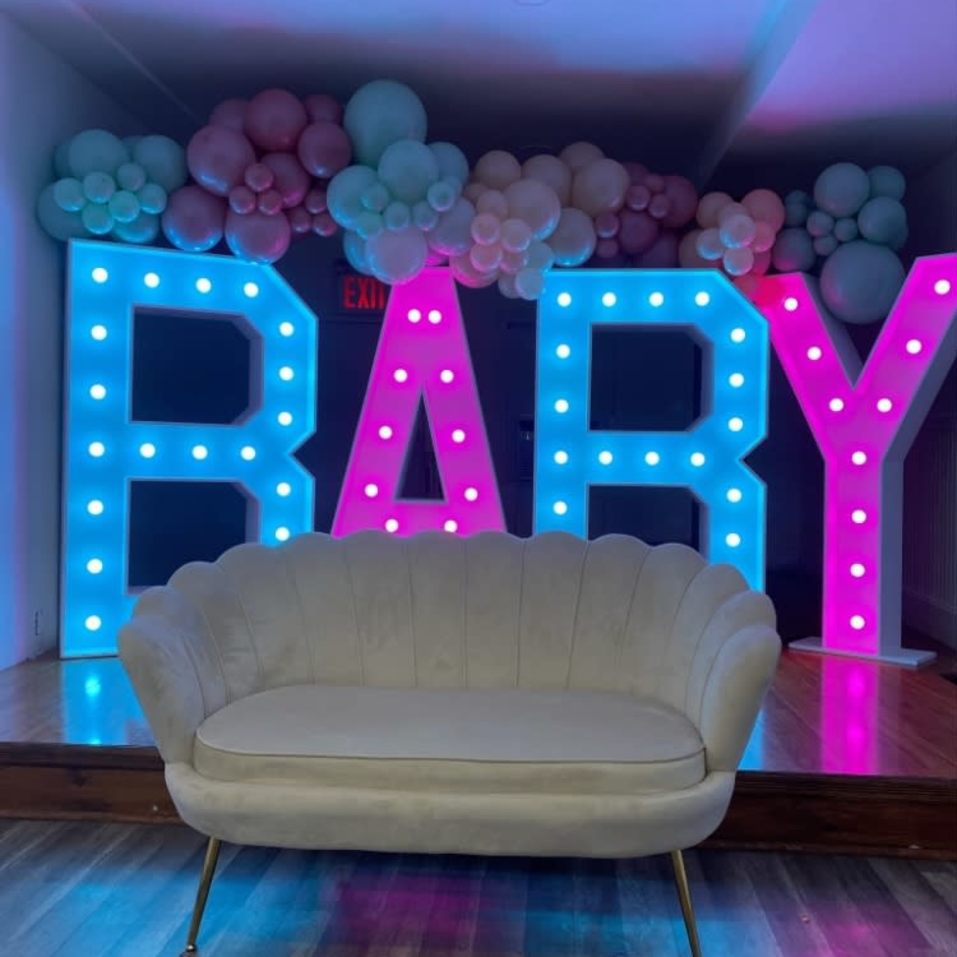 A lighted display spelling "BABY" in blue and pink is surrounded by colorful balloon arrangements, with a beige couch positioned in front of it.
