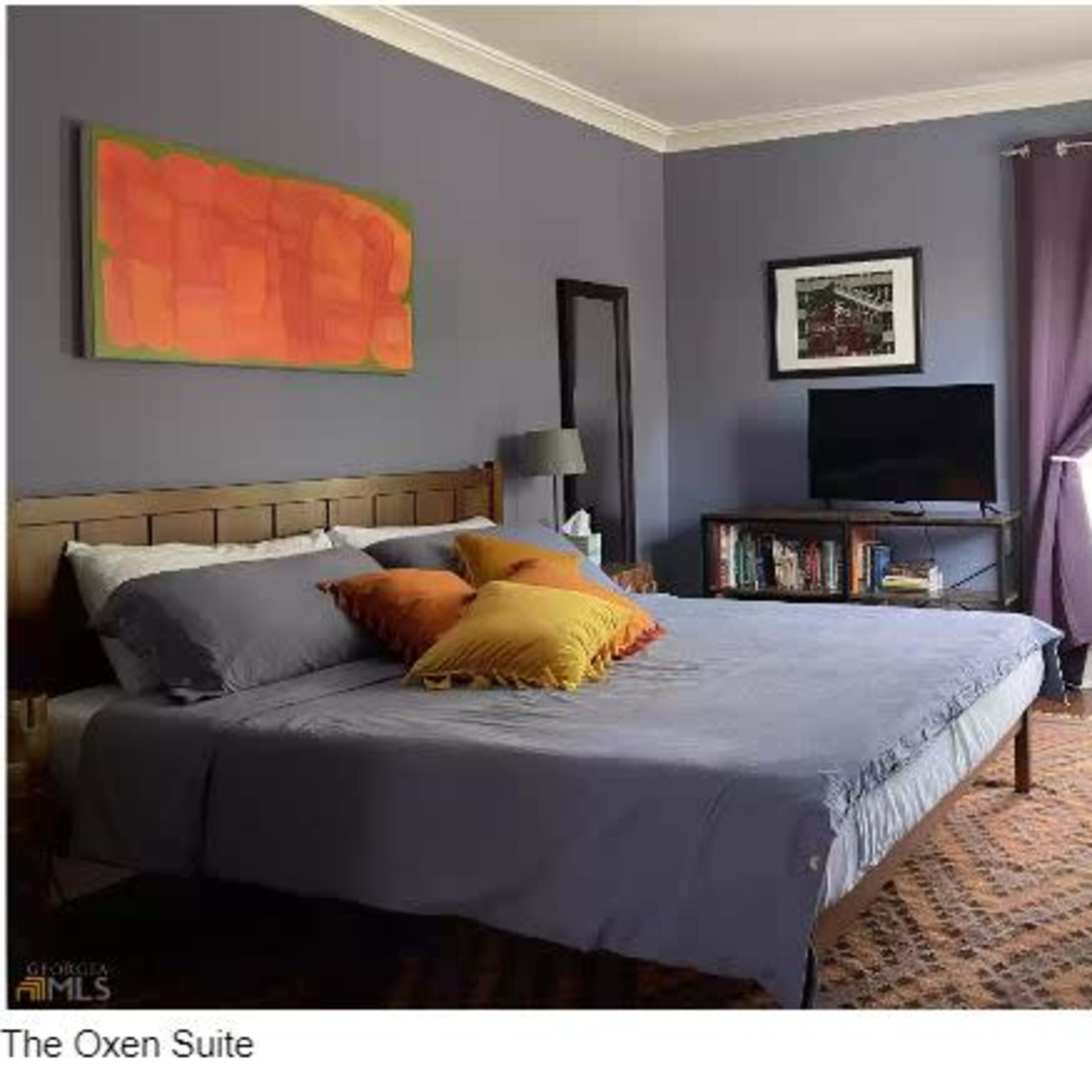 A bedroom with a bed, colorful pillows, a TV on a stand, curtains, and a window providing natural light.