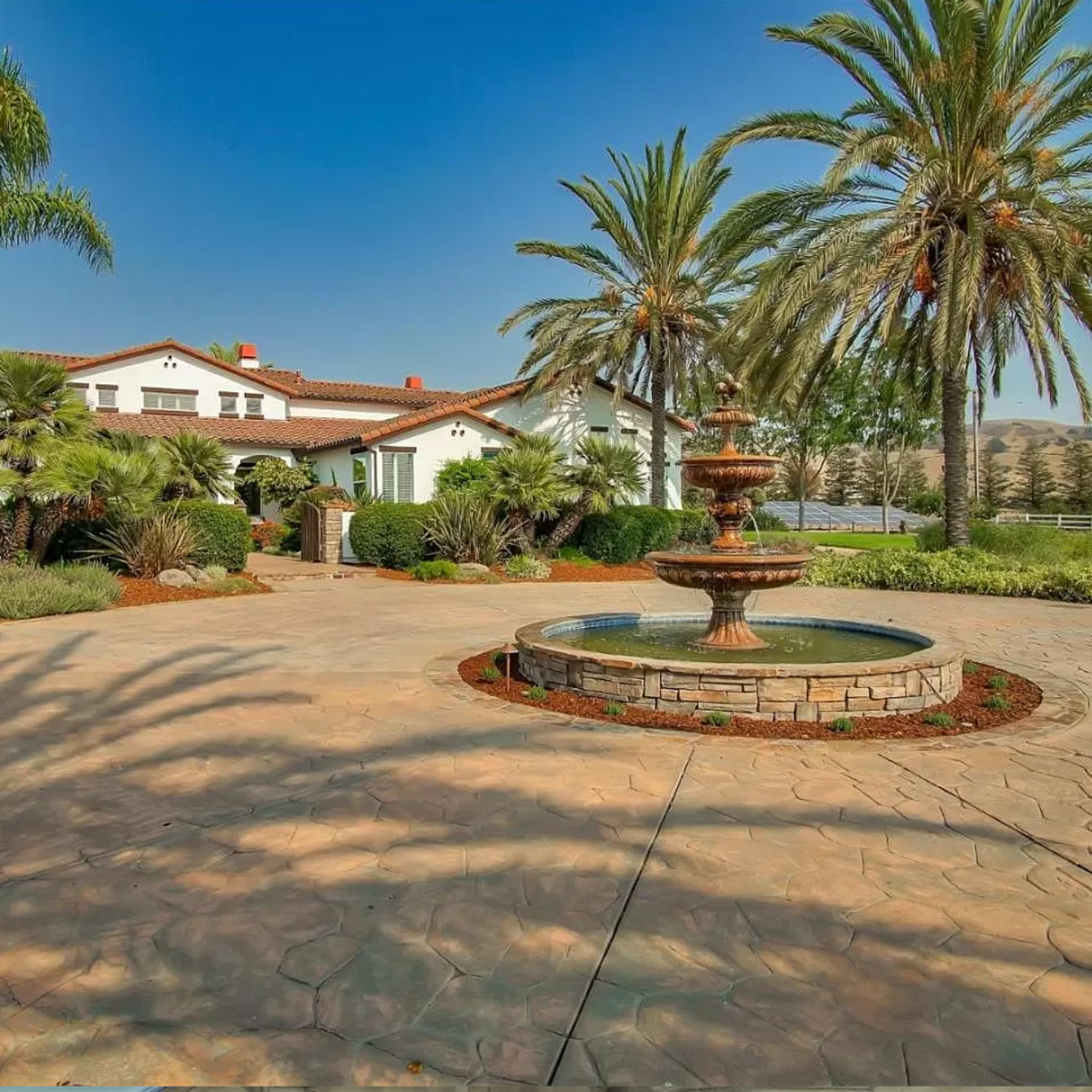 A landscaped courtyard with a fountain surrounded by palm trees and a large house in the background.