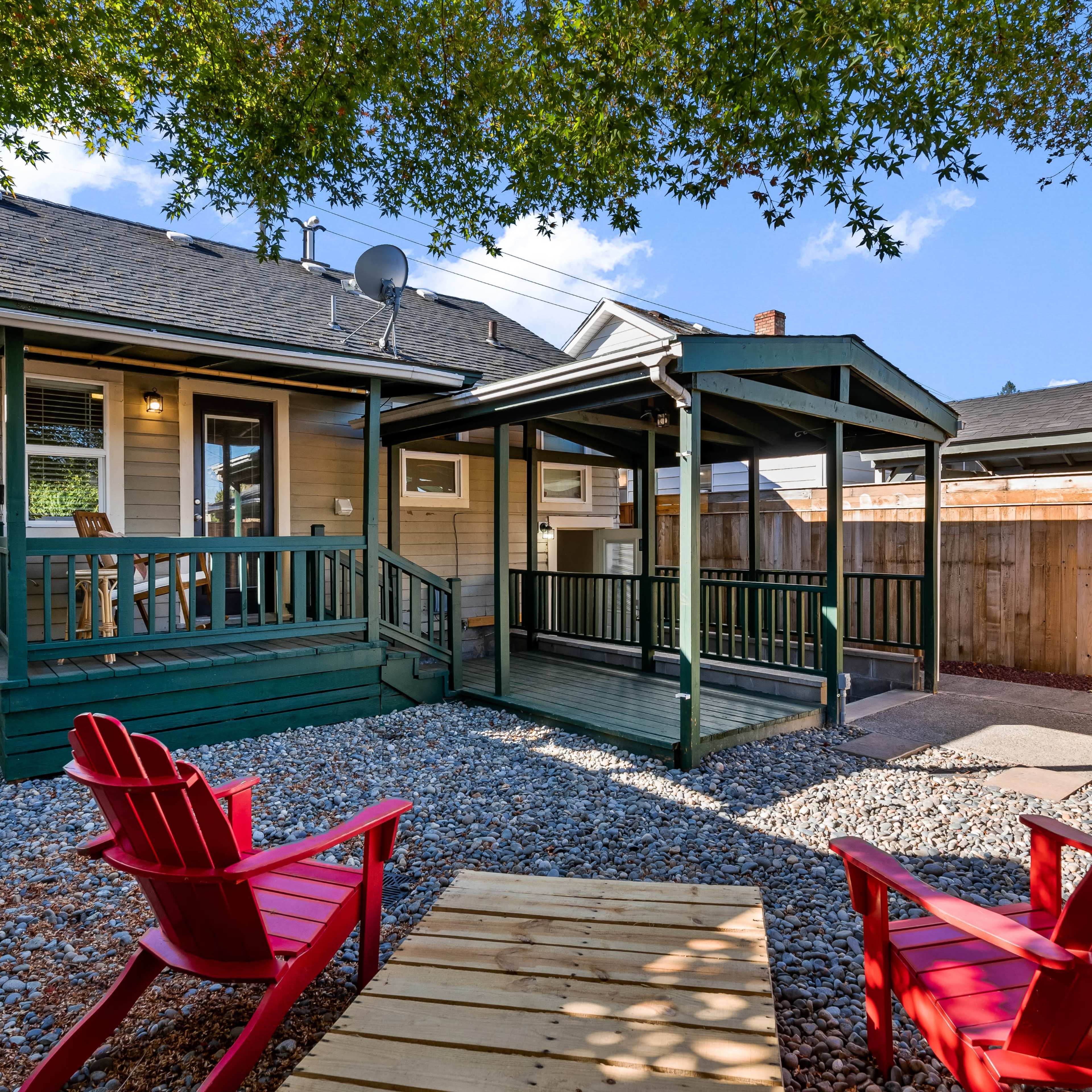 A backyard features a wooden deck with a covered seating area, surrounded by gravel and two red chairs.