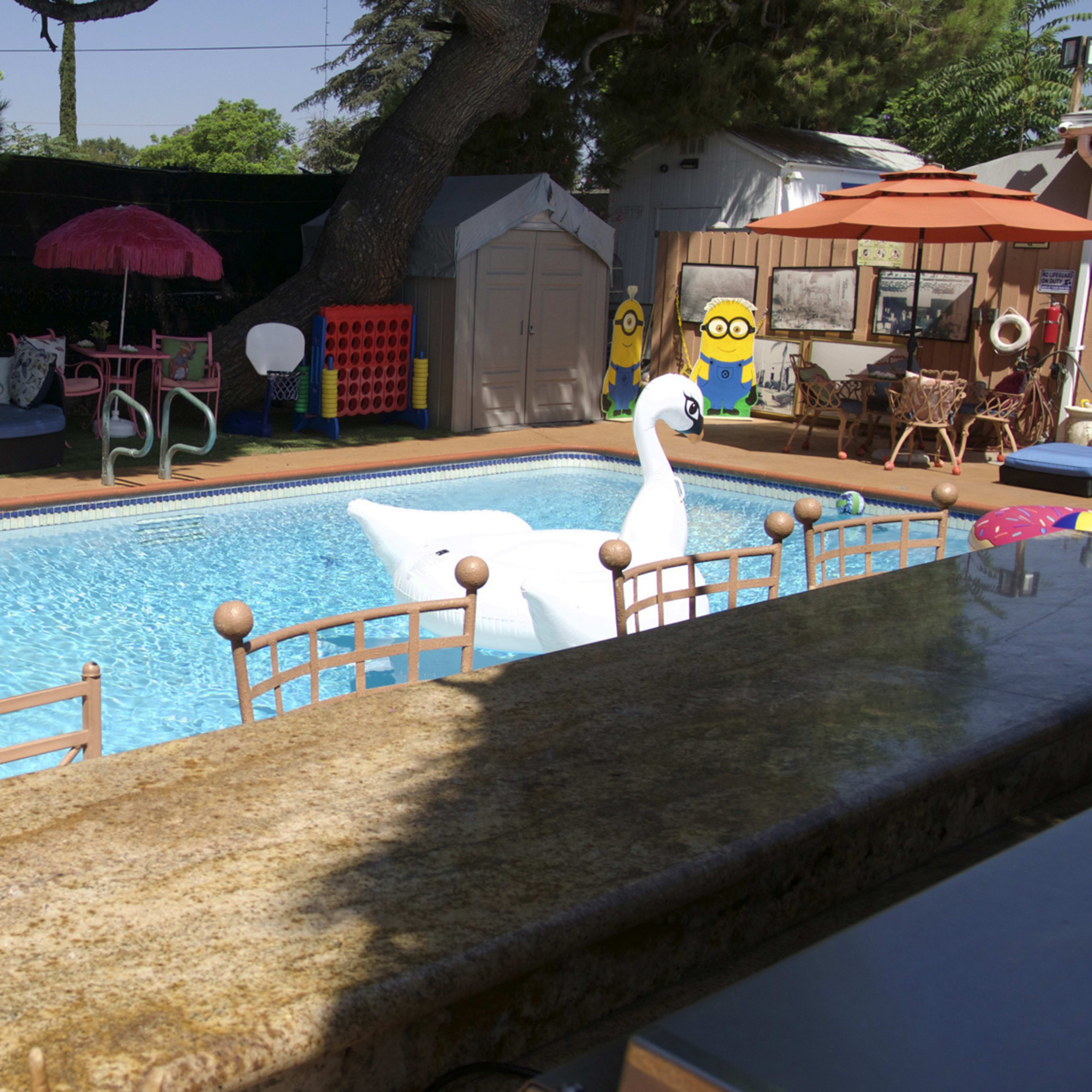 The scene features a backyard pool with inflatable yellow duck and white swan floats, surrounded by patio furniture, umbrellas, and a children's play area.