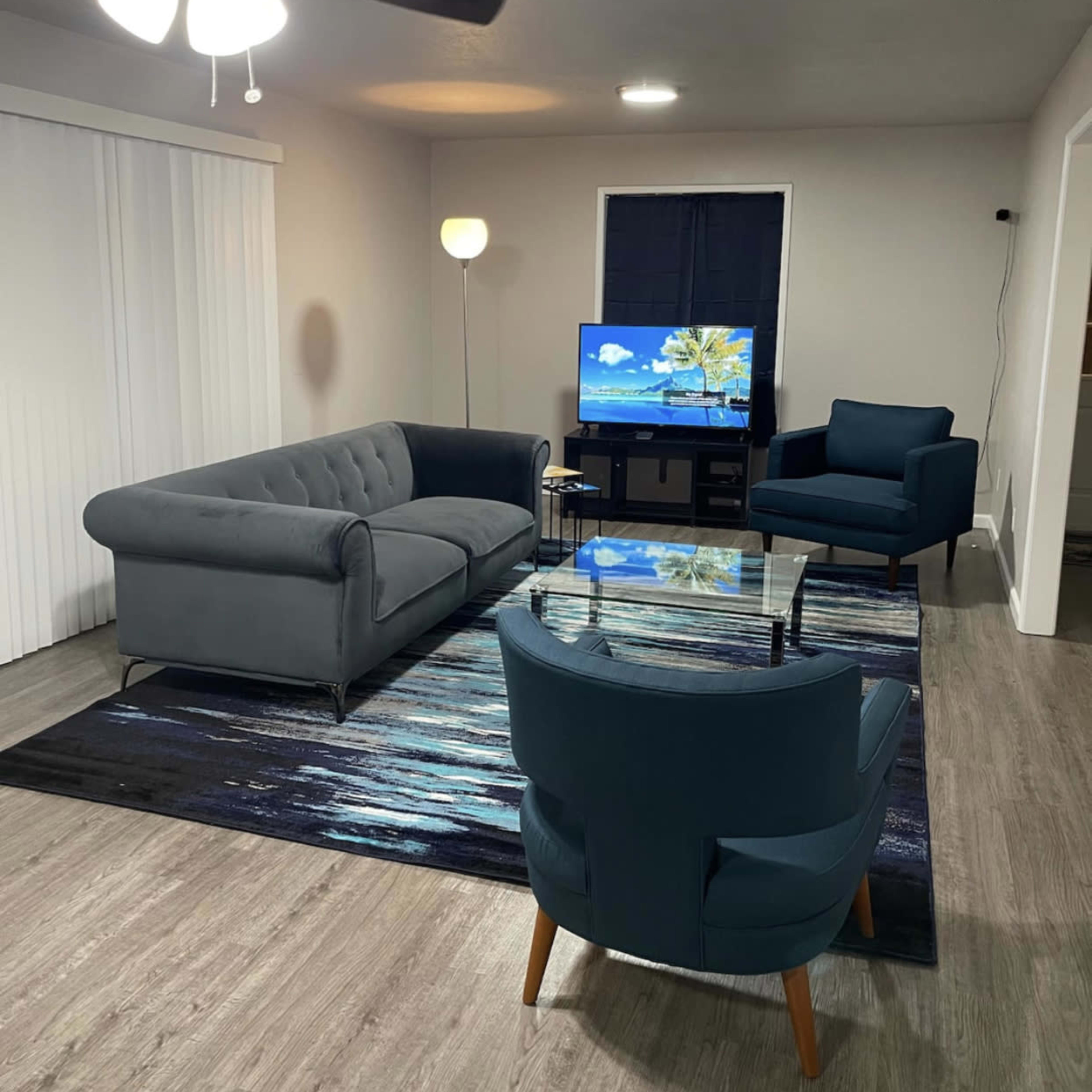 A modern living room with a gray sofa, two teal armchairs, a glass coffee table, and a television displaying a beach scene, all set on a patterned area rug.