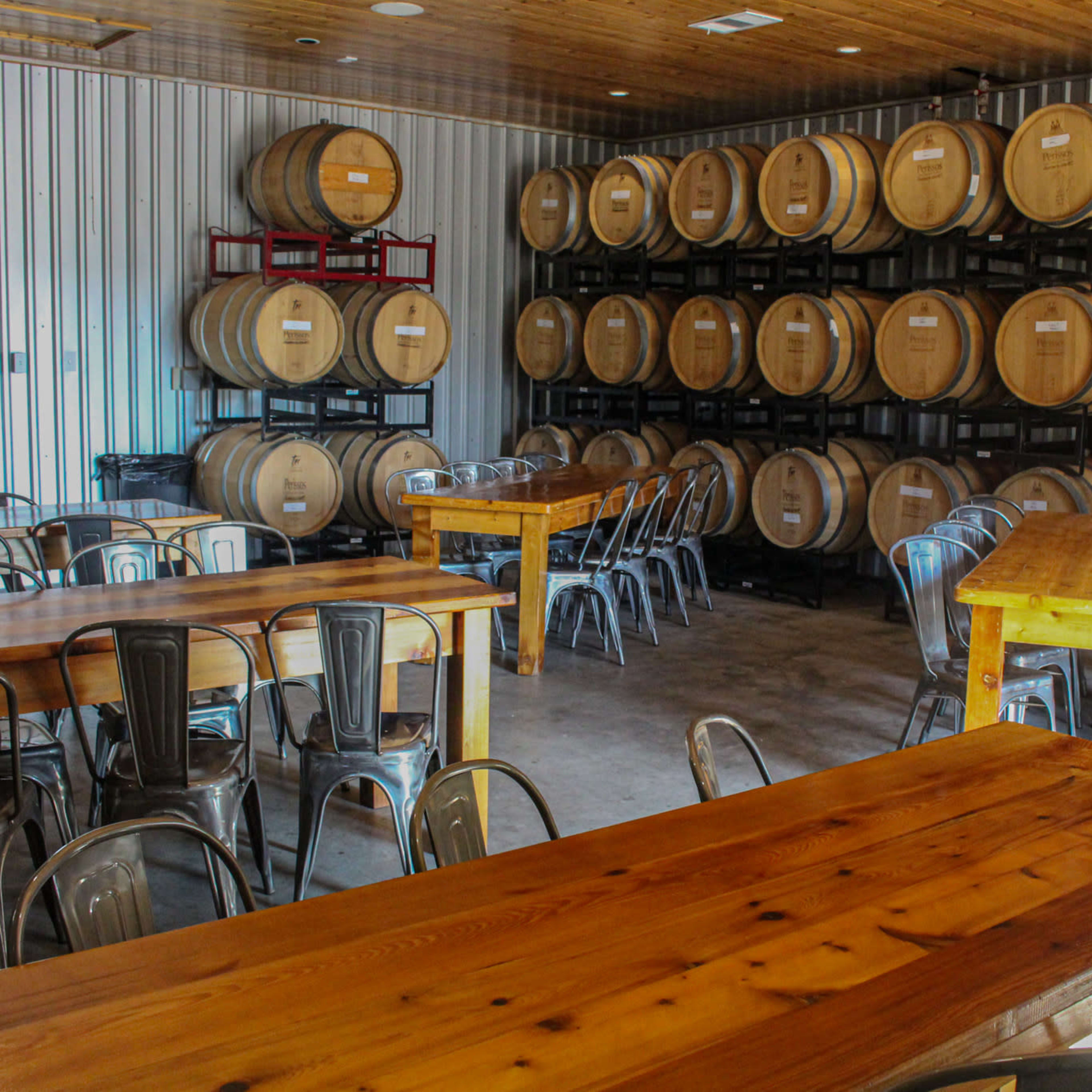 A tasting room features wooden tables and metal chairs arranged among rows of stacked wine barrels against a metallic wall.