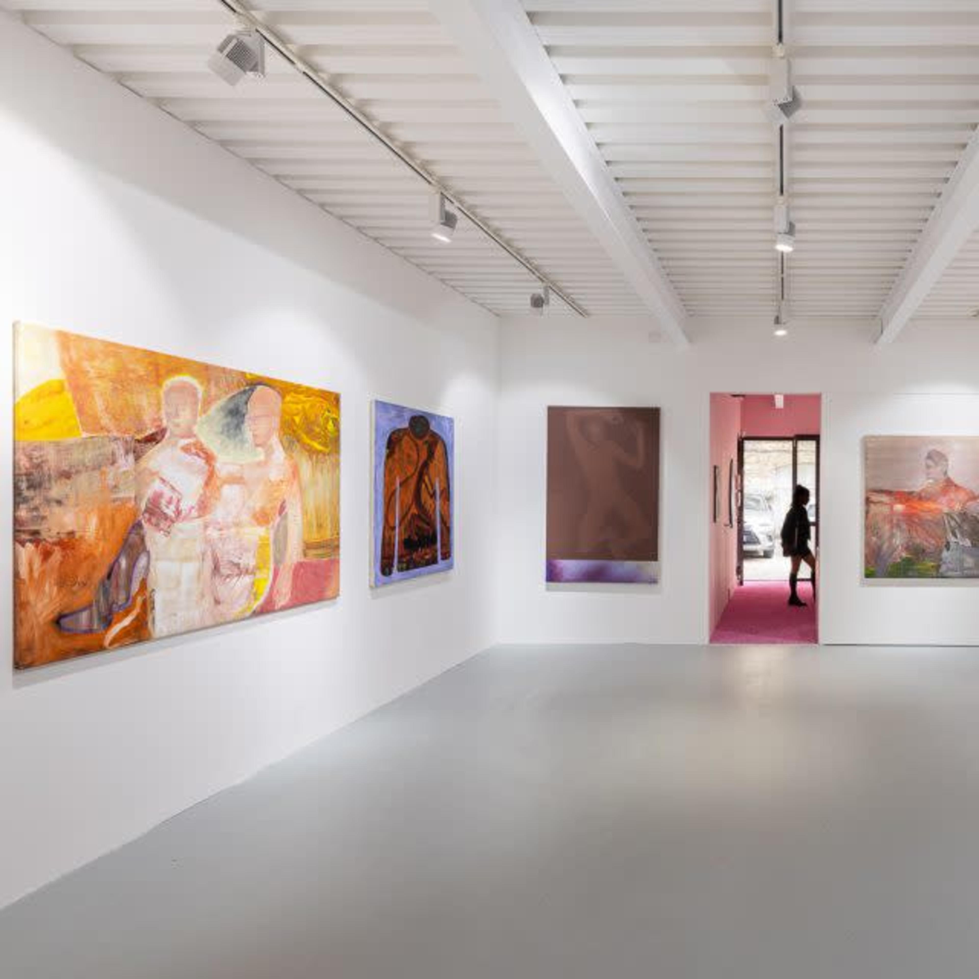 An art gallery with large, colorful paintings displayed on the white walls, and a person walking through a doorway at the far end.