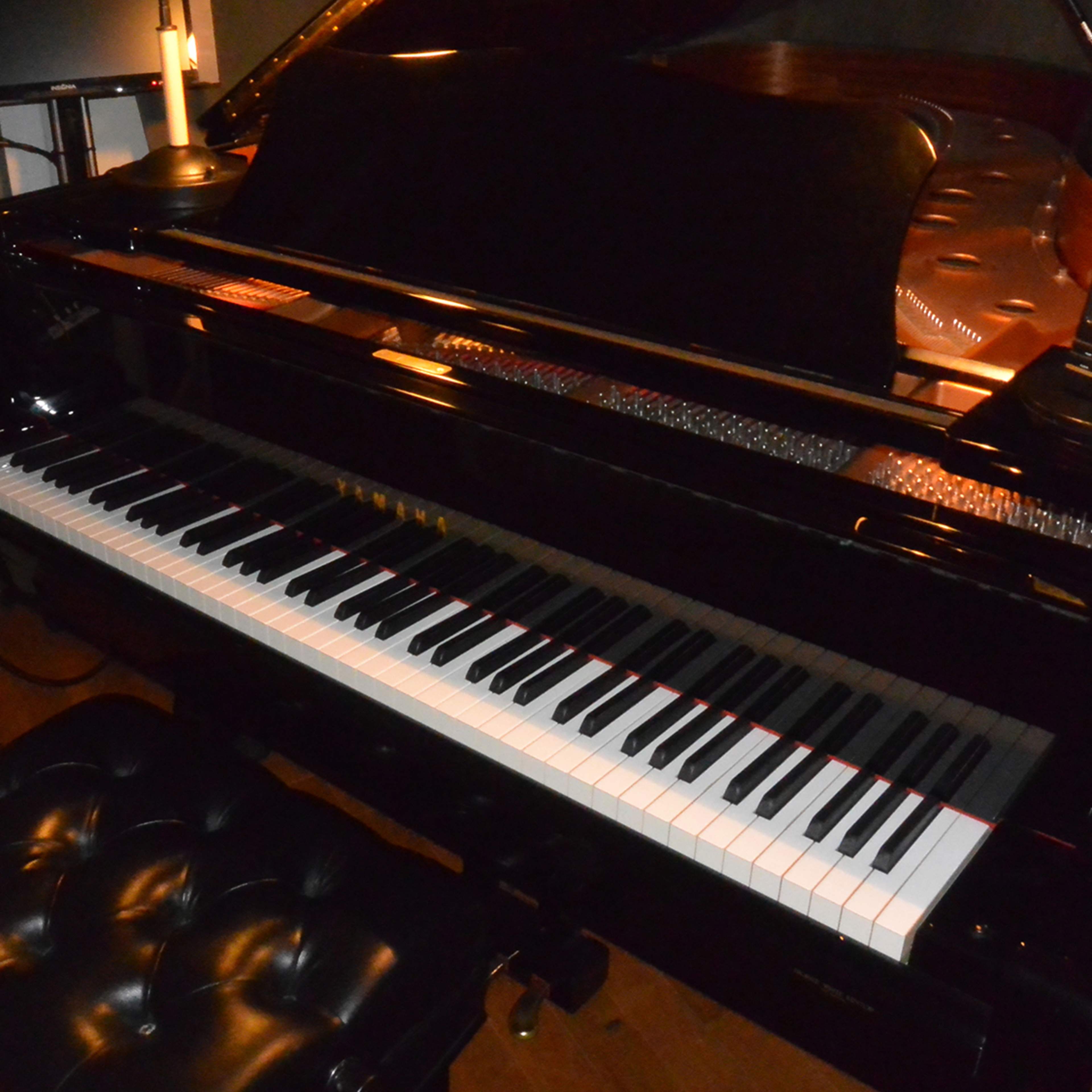 A black grand piano is positioned in a dimly lit room next to a sound mixing console and a black cushioned piano bench.