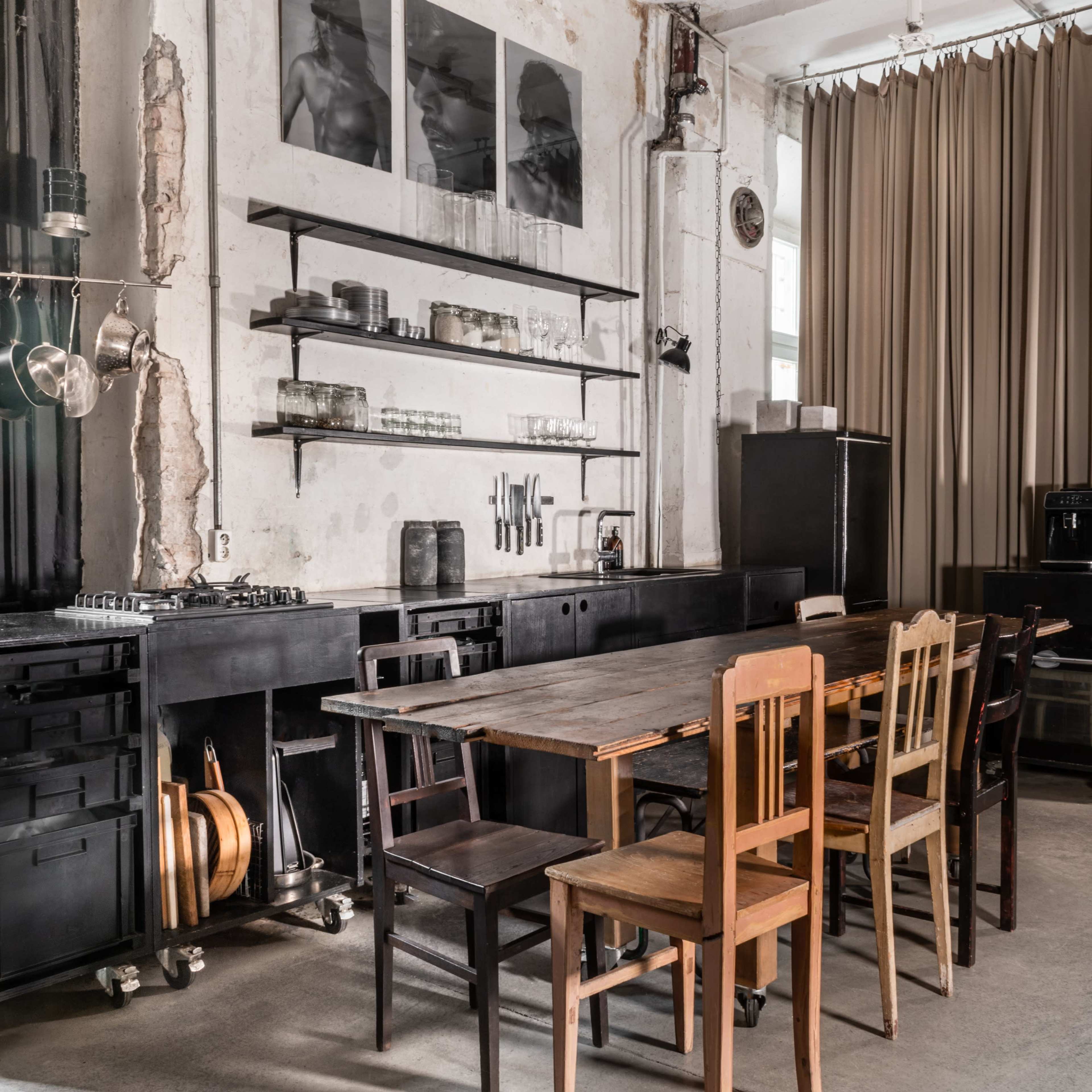 A minimalist kitchen with a long black table, various wooden chairs, and wall-mounted shelves displaying kitchenware and artwork.