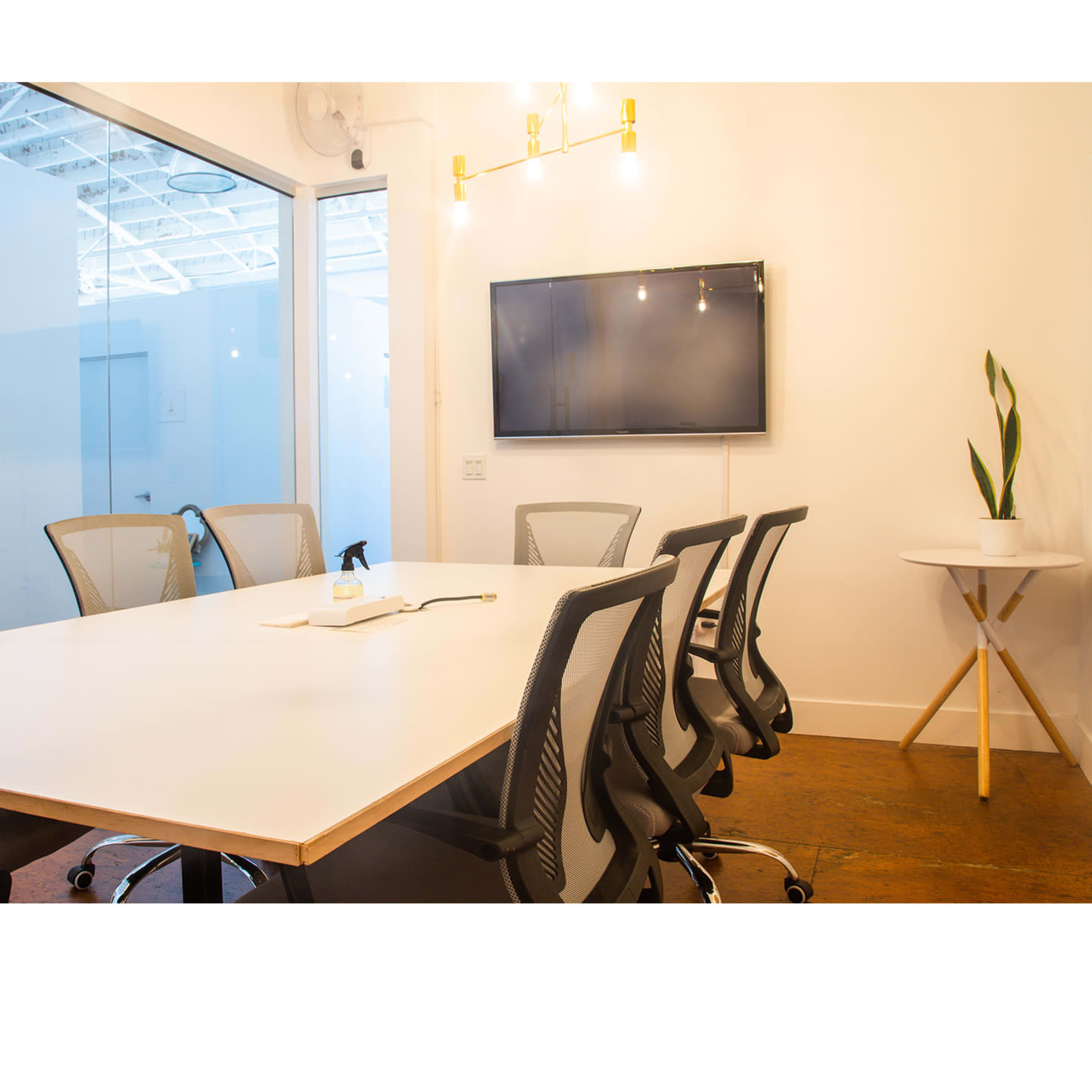 A modern conference room features a long white table surrounded by ergonomic chairs, a wall-mounted television, and a small plant on a side table.