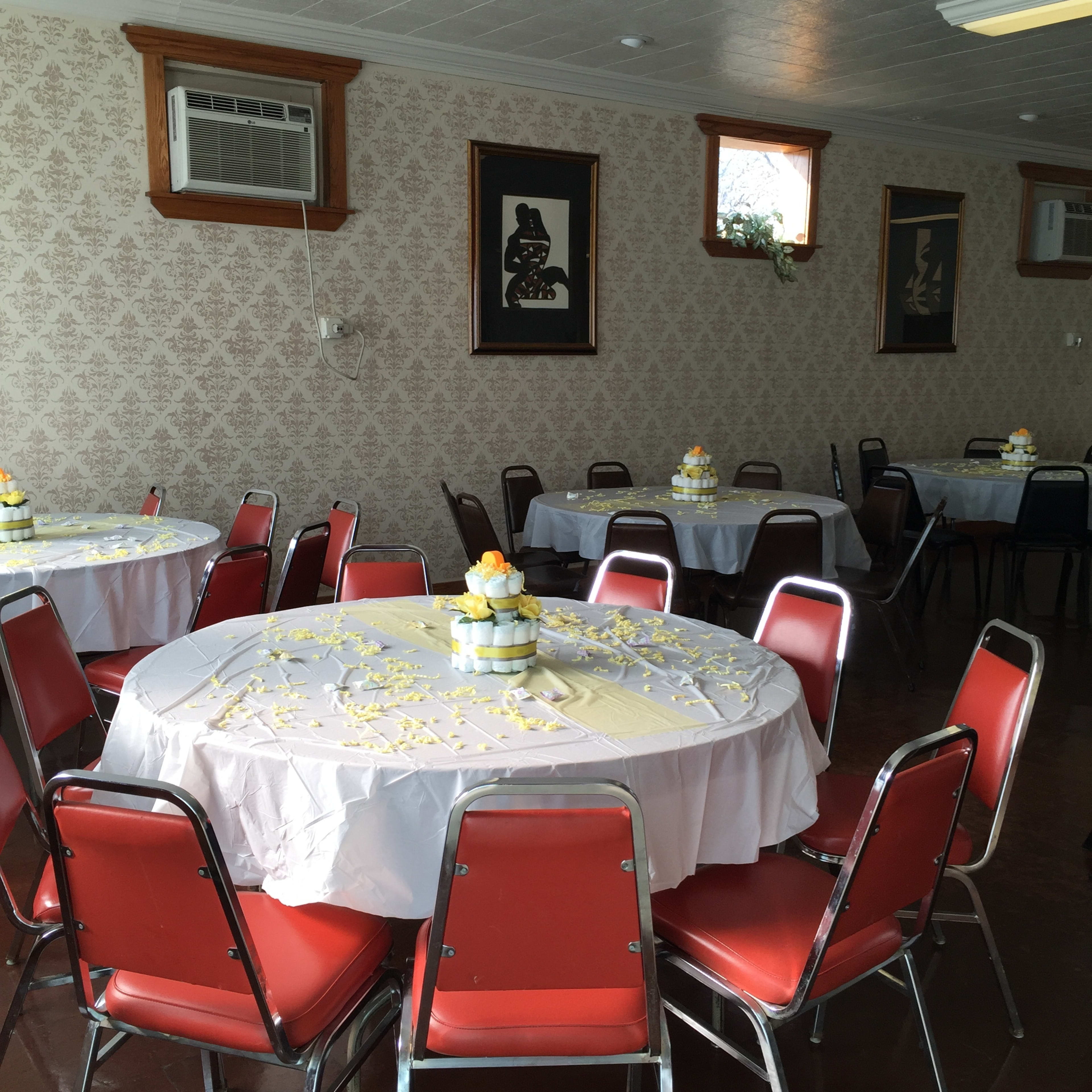 A dining hall with several round tables covered in white tablecloths and arranged with red chairs, decorated with small floral centerpieces.