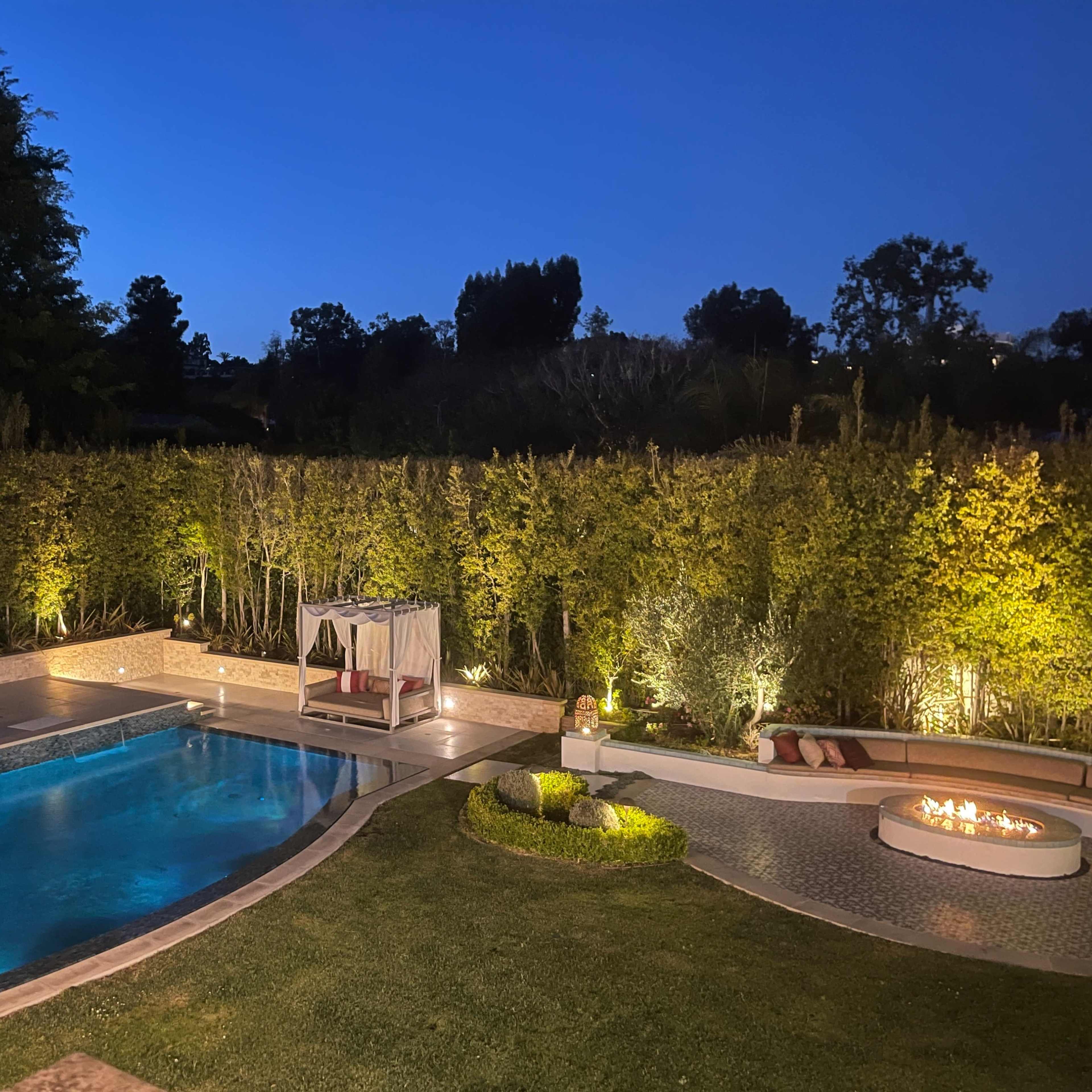 A well-lit backyard pool area surrounded by hedges, featuring a seating arrangement by the fire pit and a gazebo.
