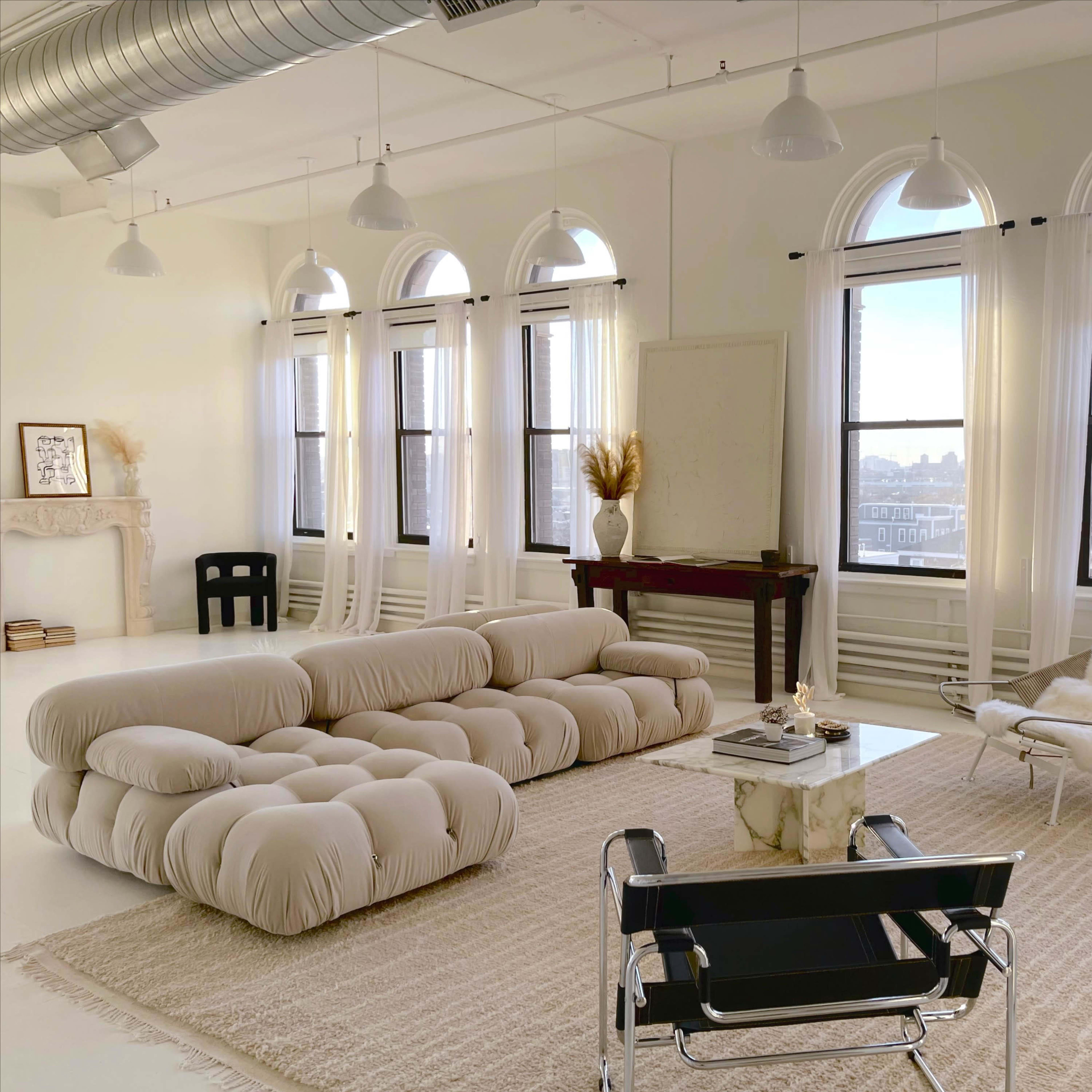 A spacious, minimalist living room featuring a plush beige sofa, large windows with sheer curtains, and a simple coffee table surrounded by modern chairs.