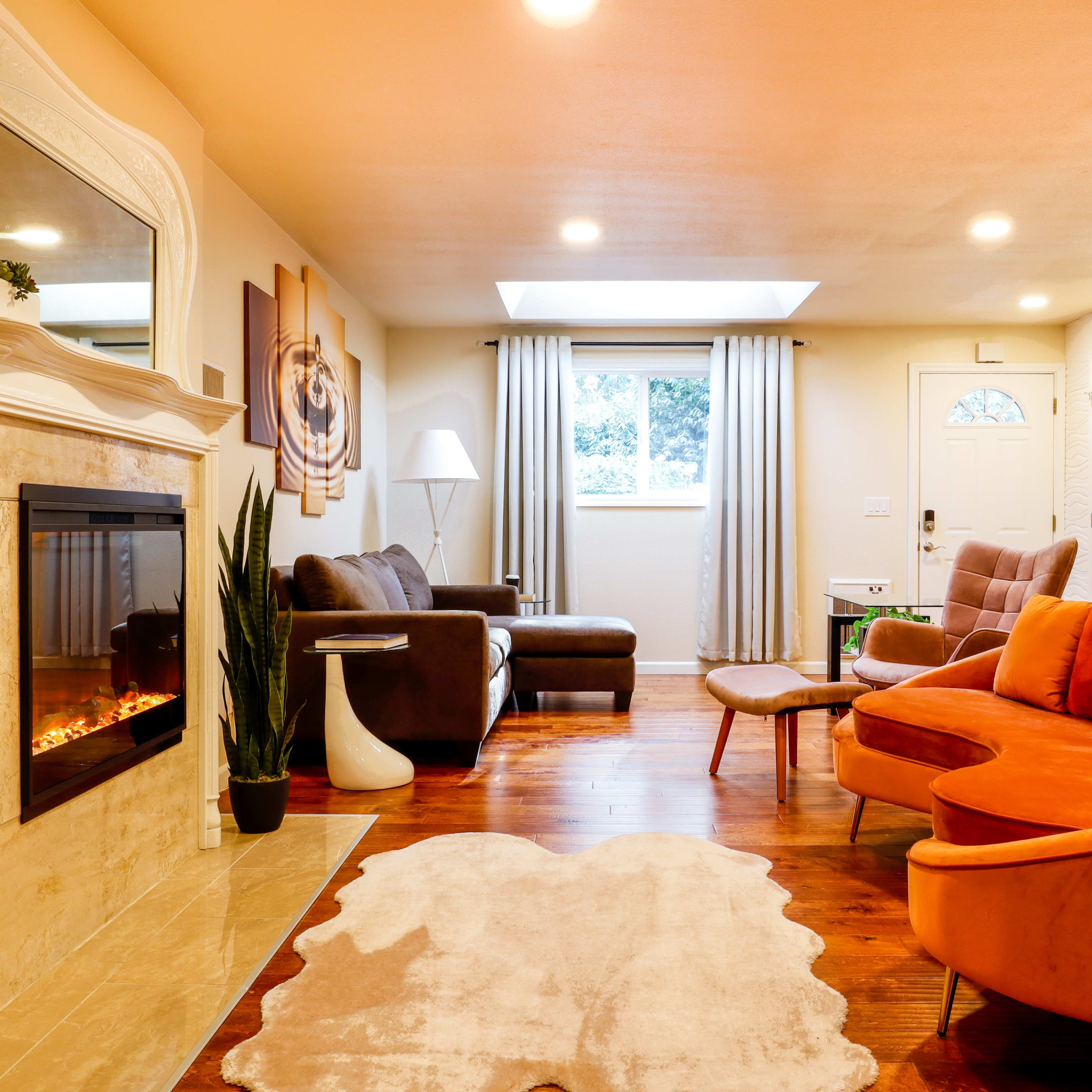 A cozy living room featuring a fireplace, a large orange sectional sofa, a brown couch, and a light-colored area rug on hardwood floors.