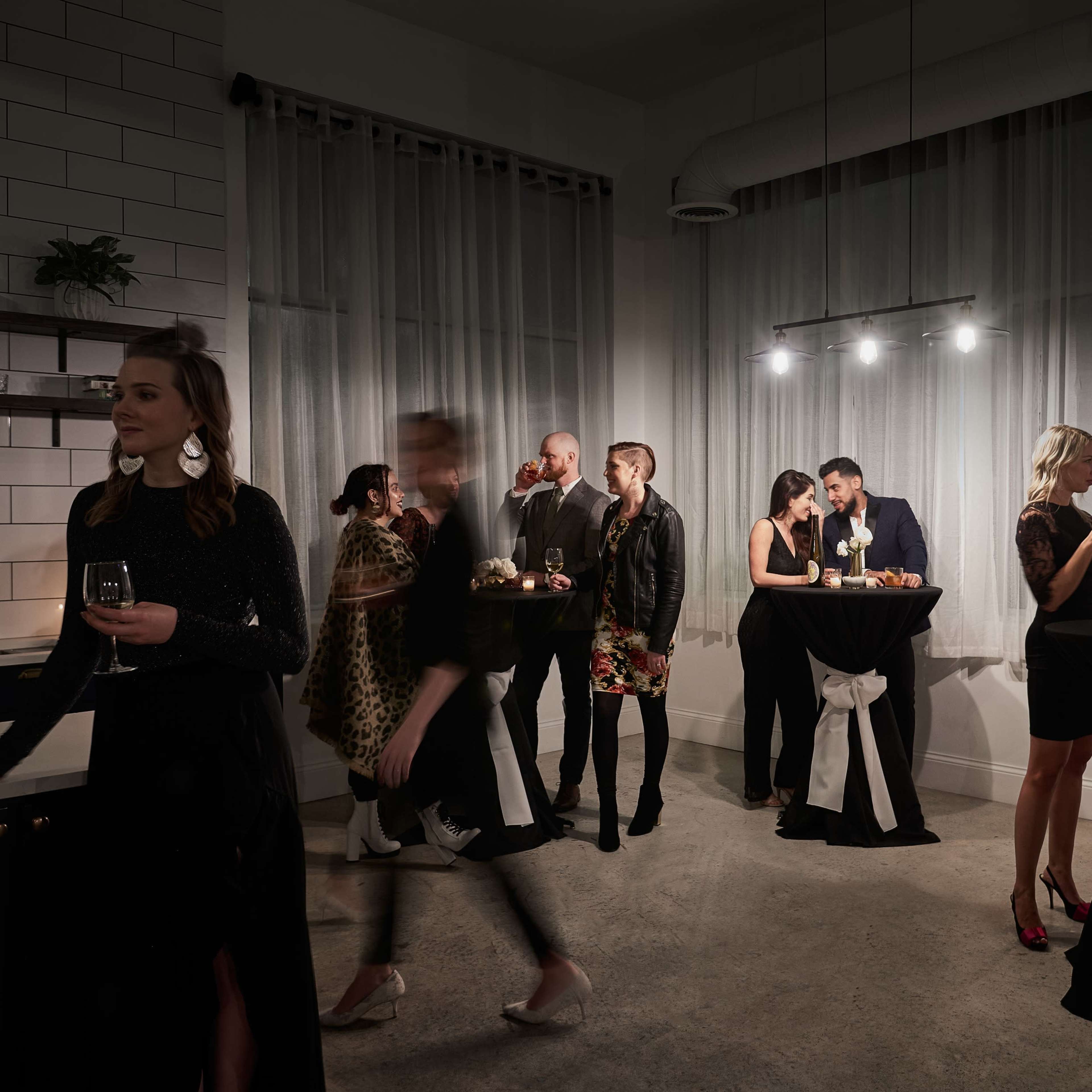 A group of elegantly dressed people socialize at a dimly lit upscale event, while others engage in conversation near tall tables adorned with flowers and candles.