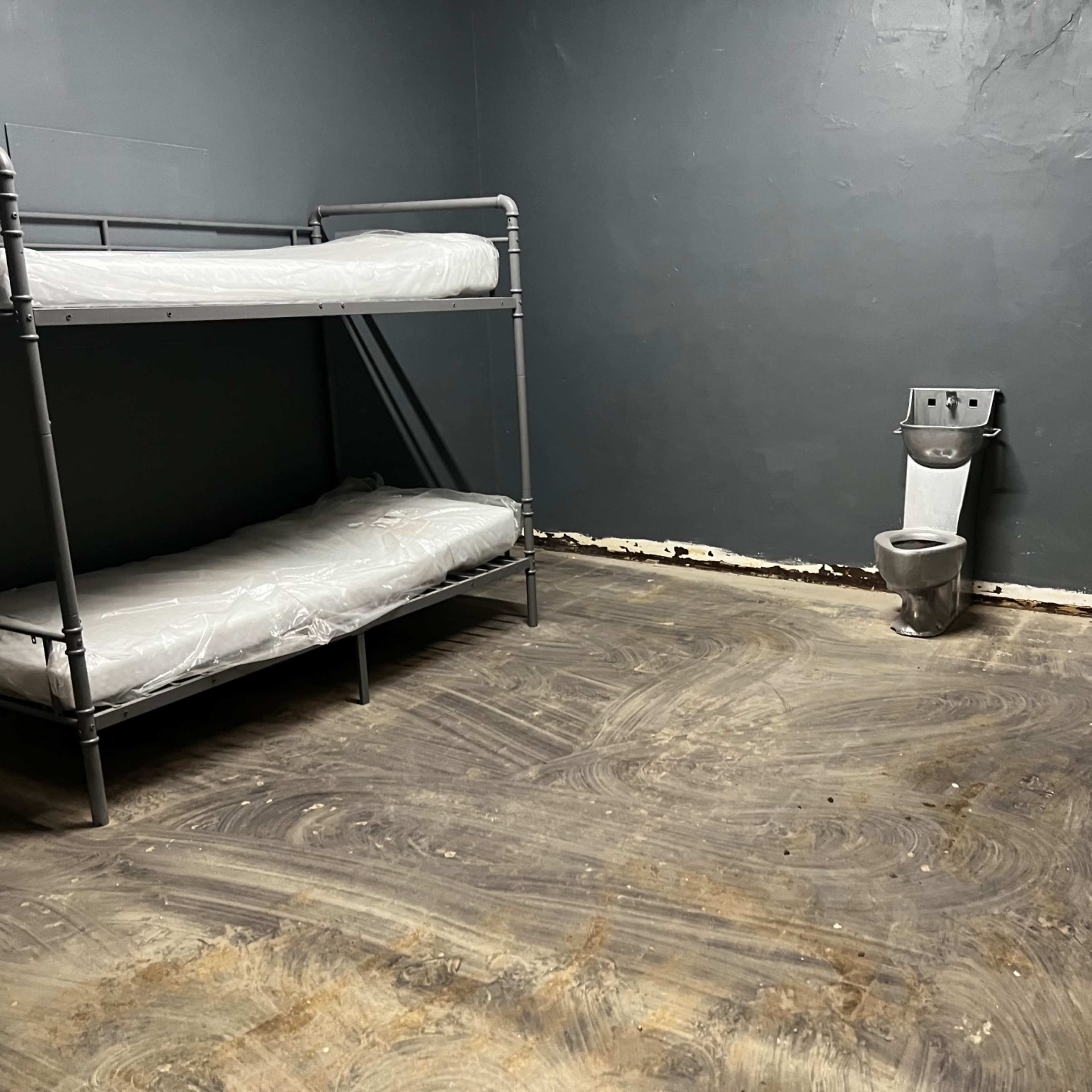 A sparse room with a bunk bed on one side, a single empty bed on the floor, a toilet in the corner, and a gray storage unit against the wall.