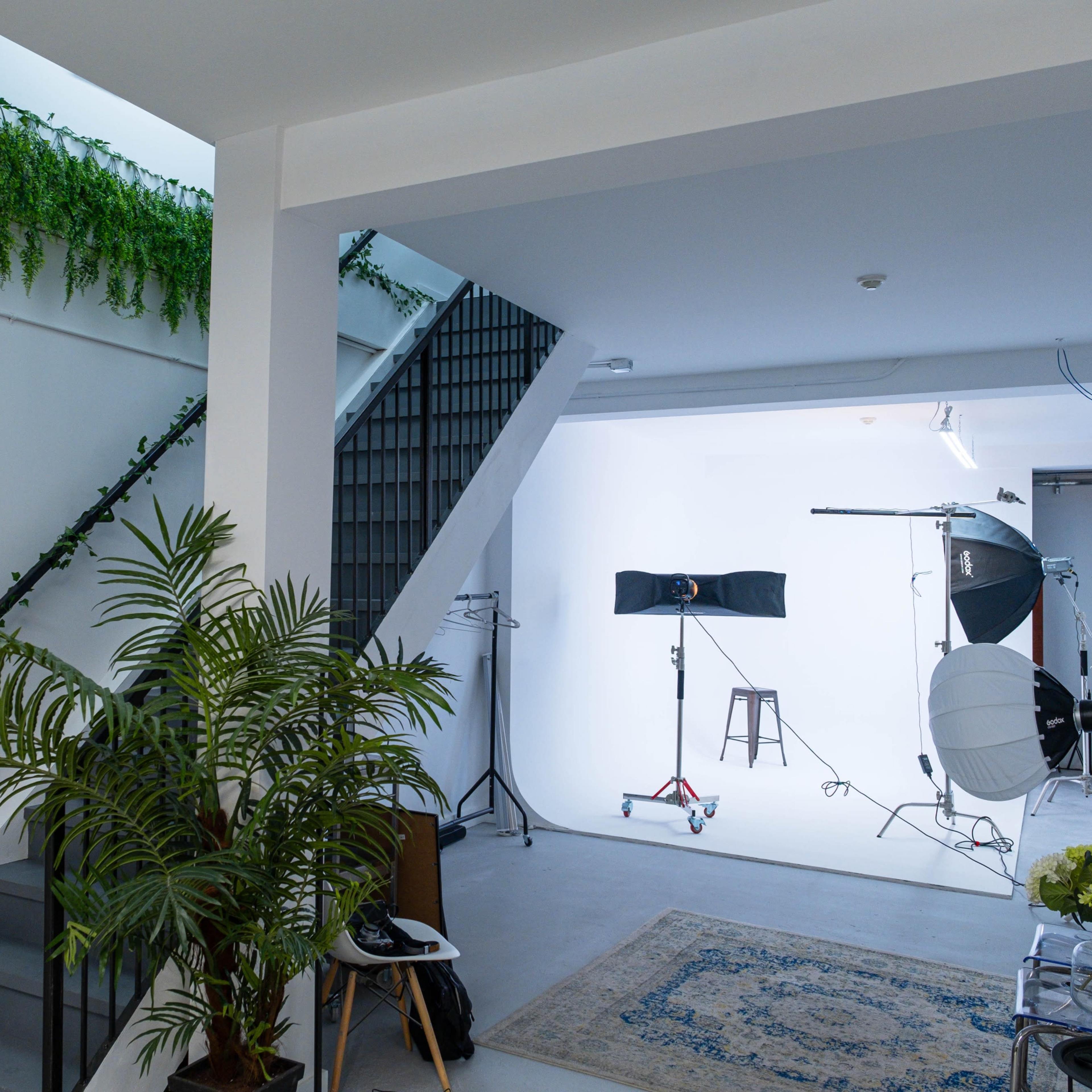 A well-lit photography studio with a staircase, plants along the walls, and lighting equipment set up for a photoshoot.