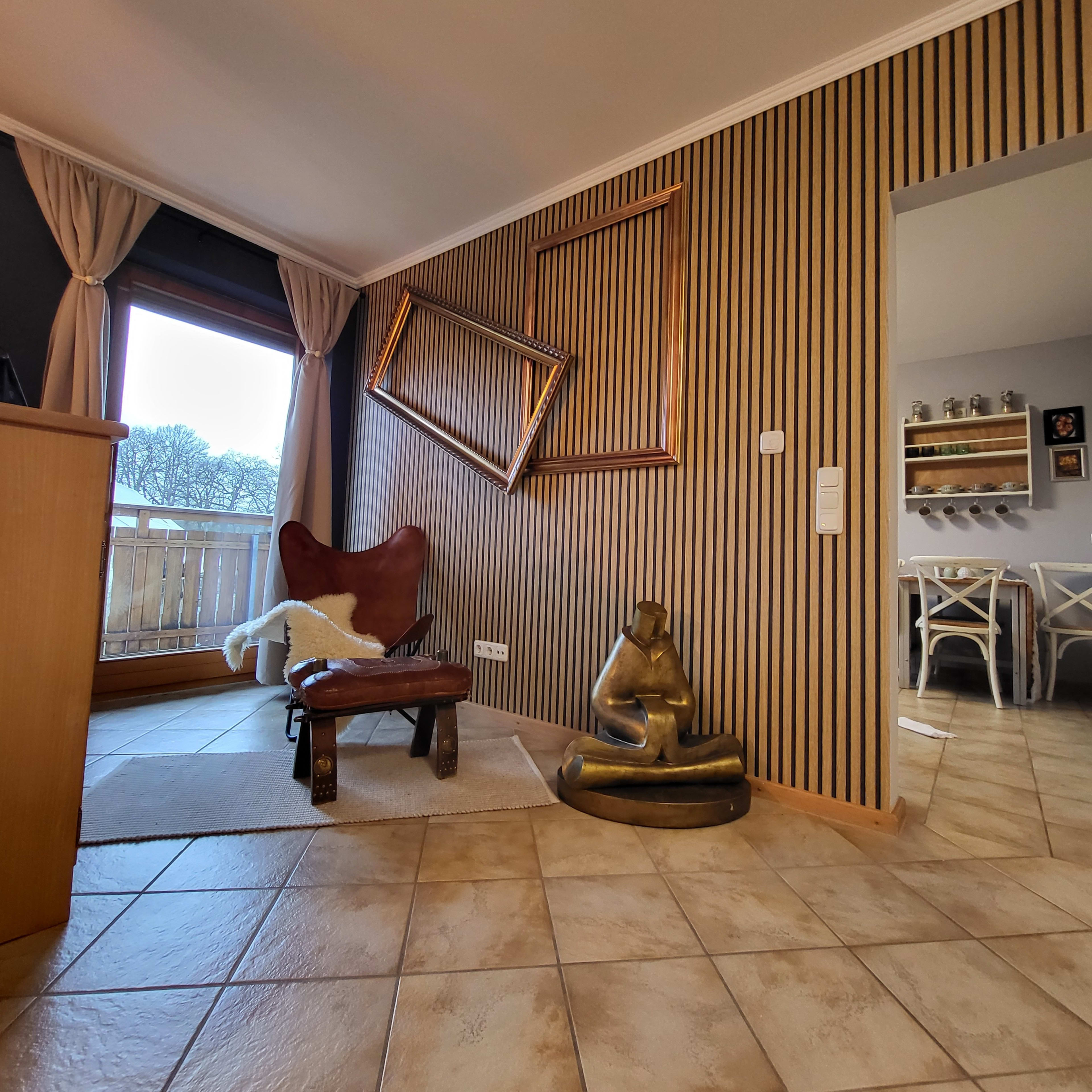 A corner of a room with a wooden floor, a leather chair, a statue of a seated figure, and a tilted picture frame mounted on a striped wall.