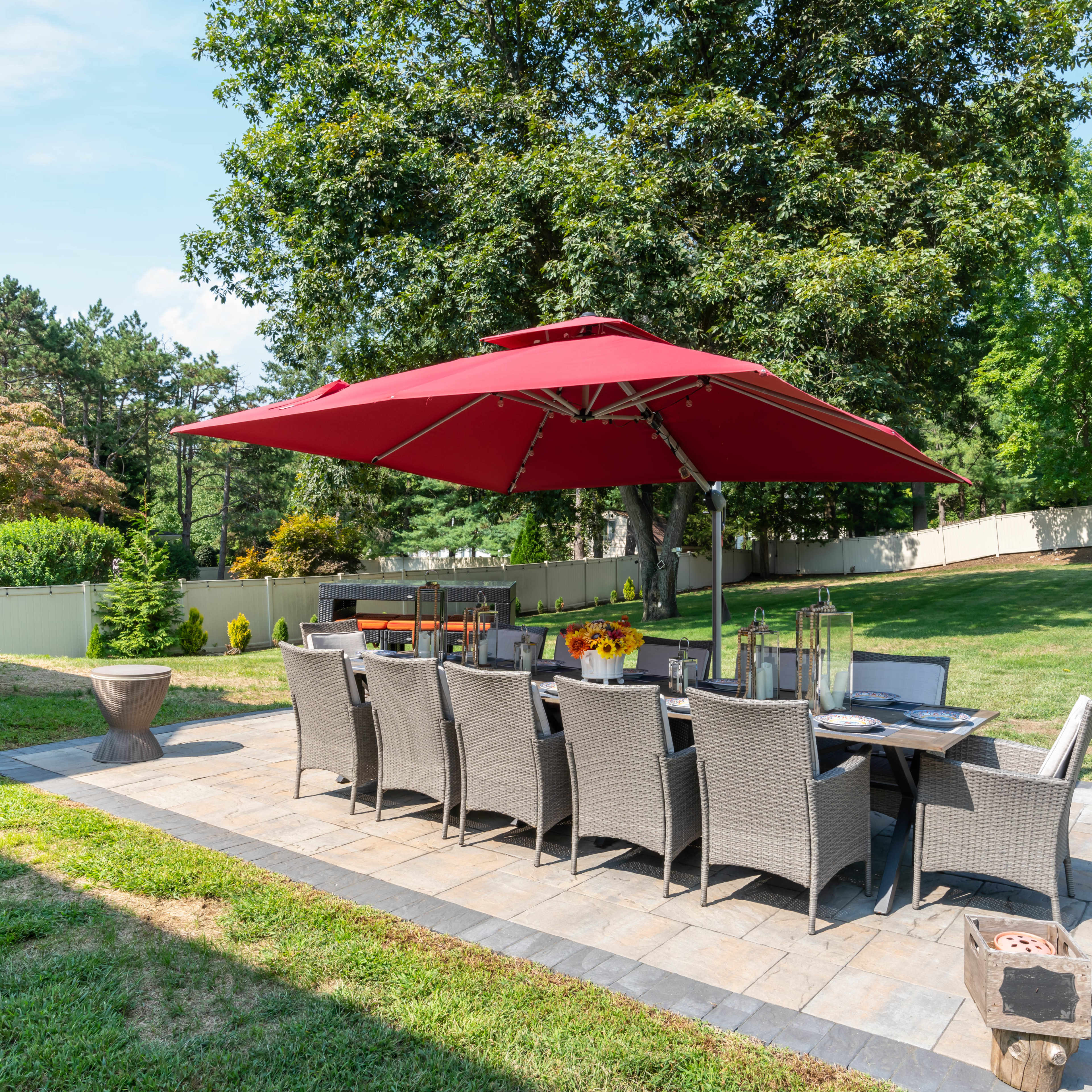 A patio dining area with a large red umbrella and a table set for ten is situated on stone pavers in a green backyard.