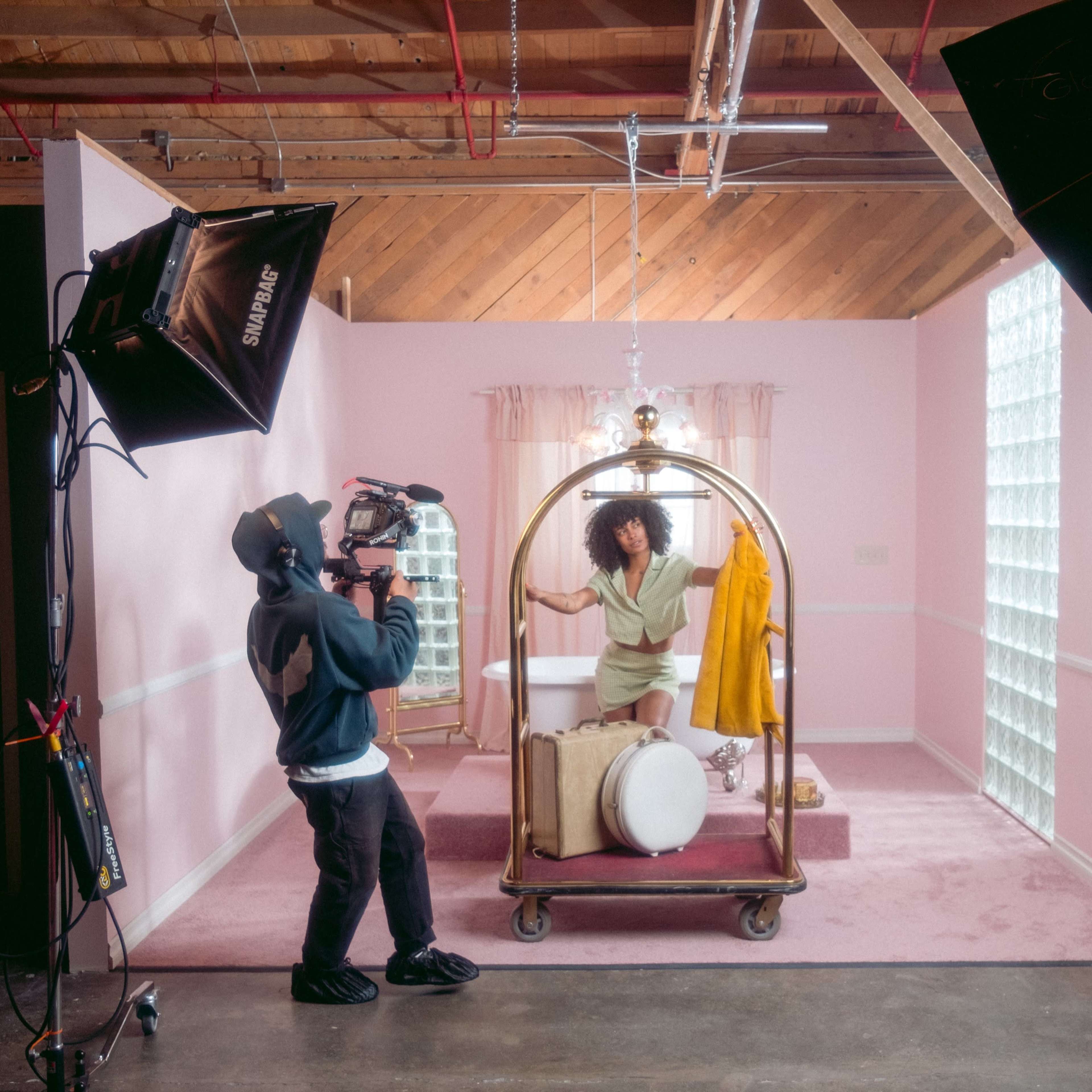 A woman poses inside a vintage bathtub on a pink set while a cameraman captures the scene with a professional camera.