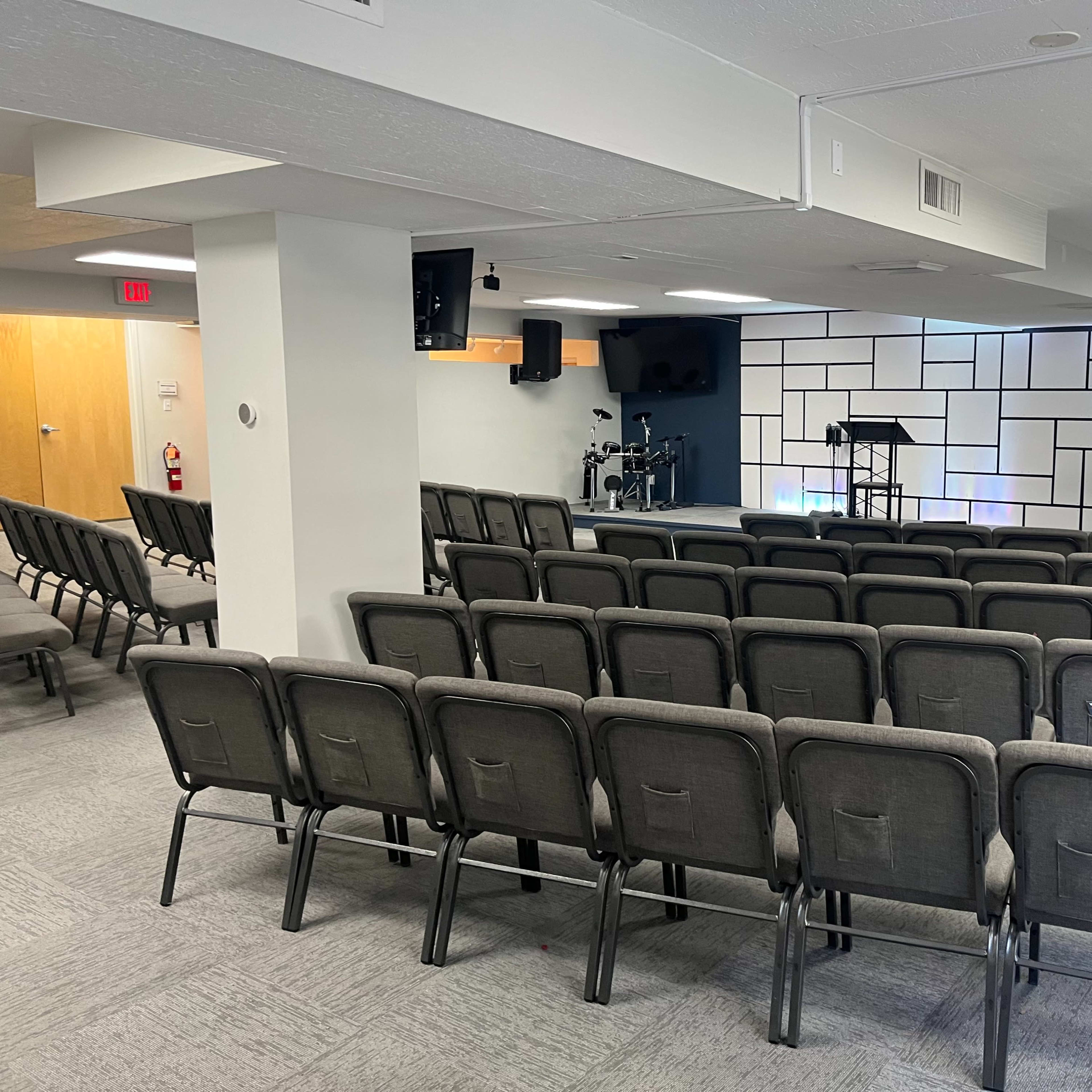 A seating area in a room with rows of gray chairs facing a small stage equipped with microphones and instruments.