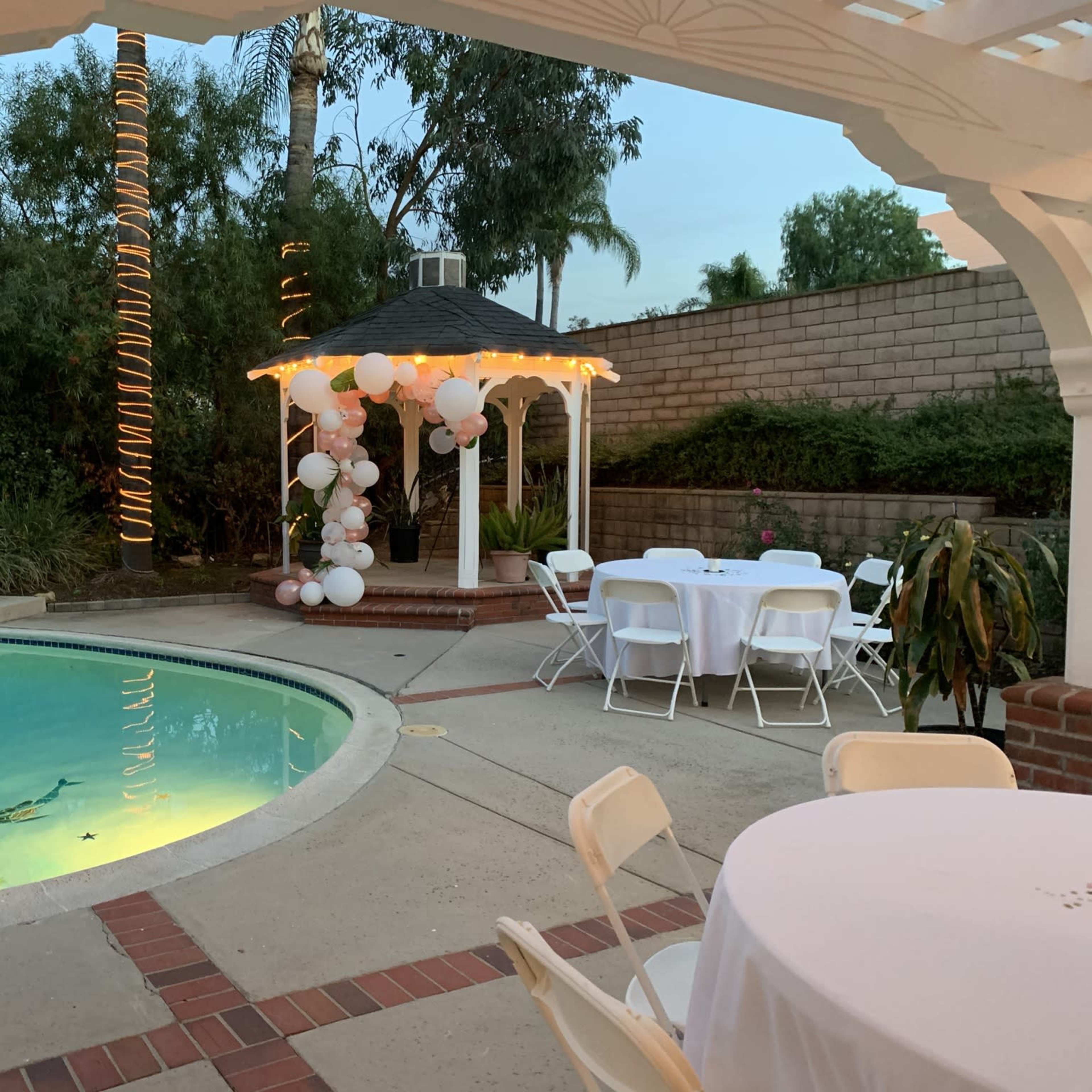 A gazebo adorned with balloons is set up near a pool and tables for an outdoor gathering in a landscaped backyard.