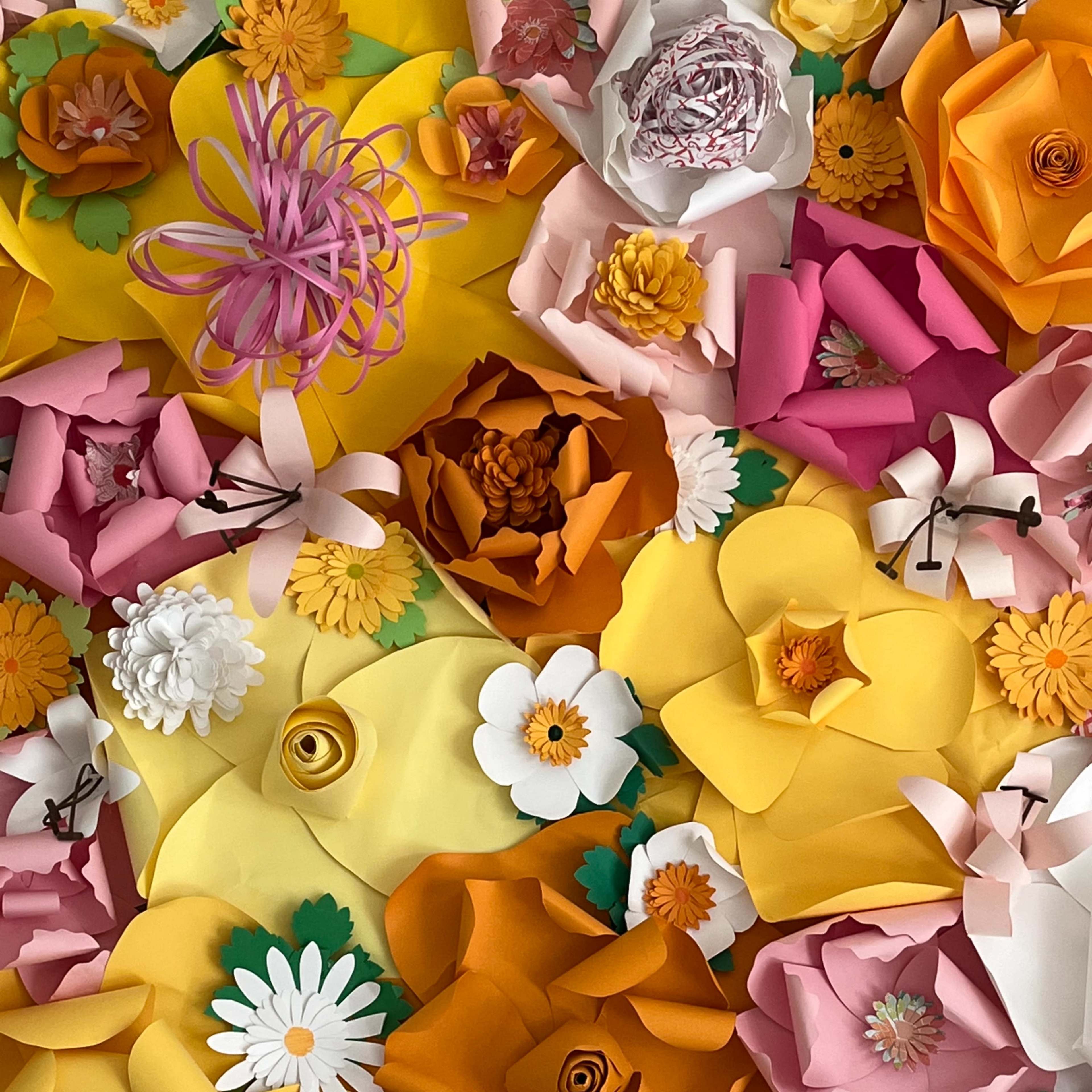A vibrant arrangement of various paper flowers in shades of yellow, orange, pink, and white, set against a colorful backdrop.