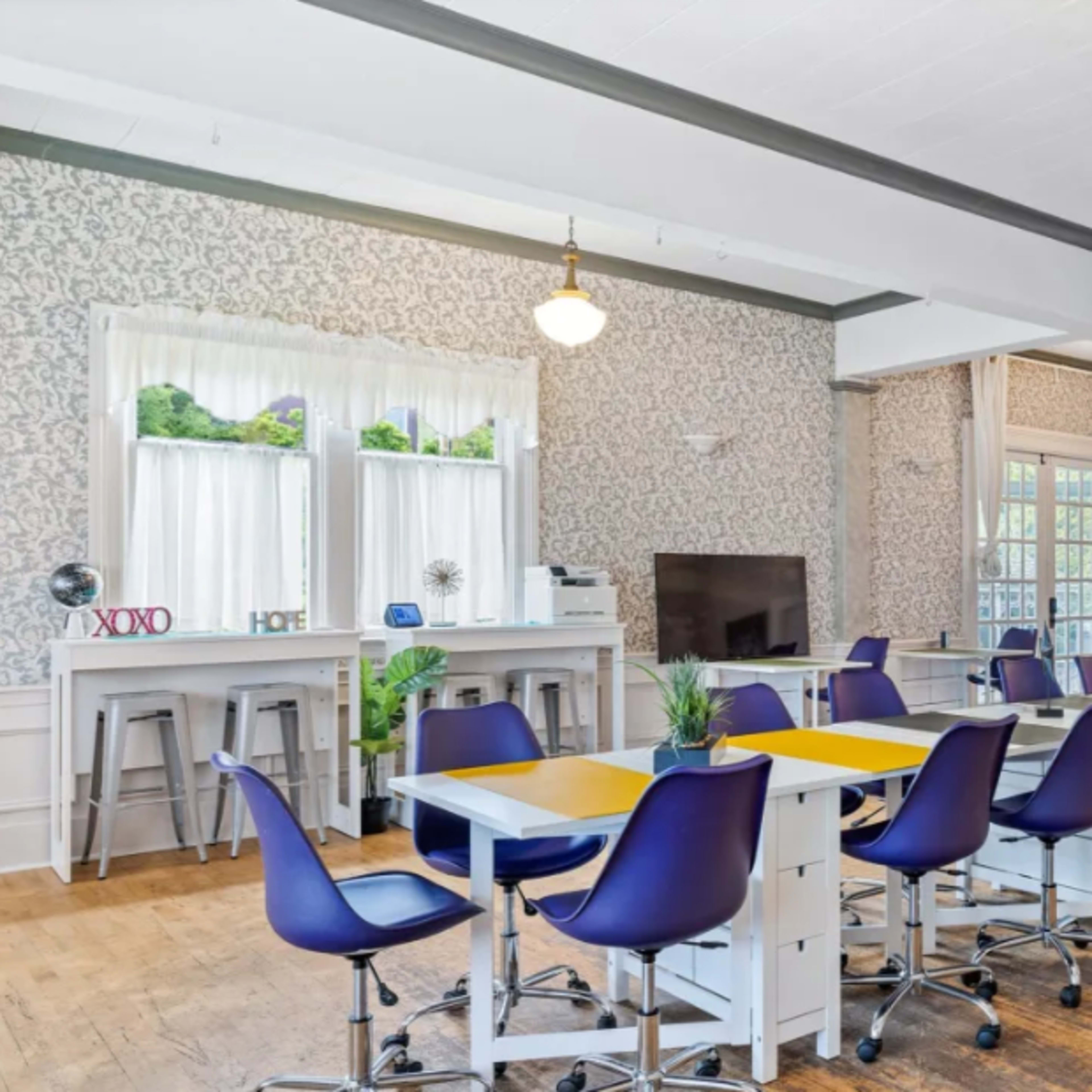 A spacious room with several purple chairs around yellow tables, large windows, and lights illuminating the space.